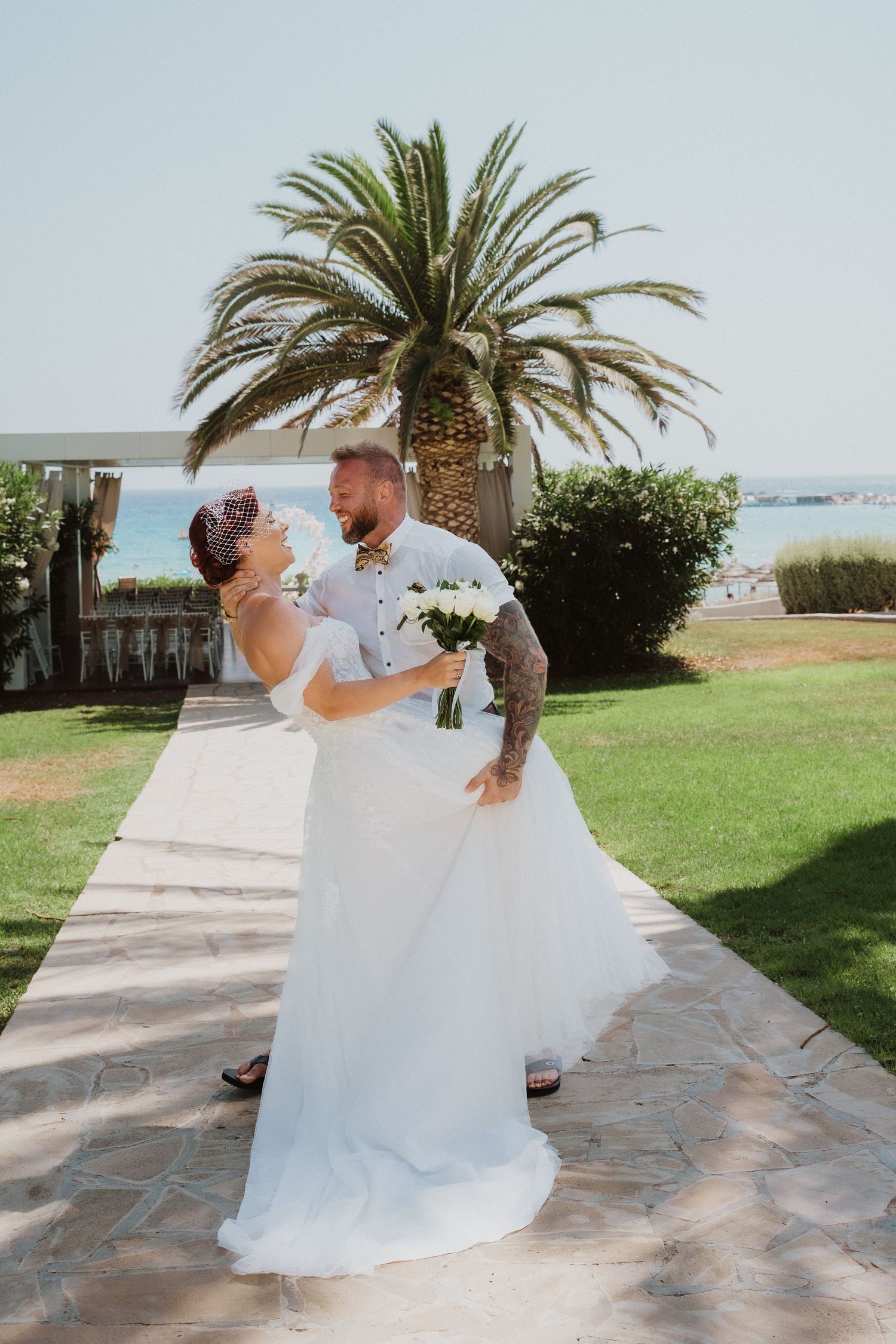 Romantic wedding couple shares intimate moment on coastal pathway with palm tree backdrop.