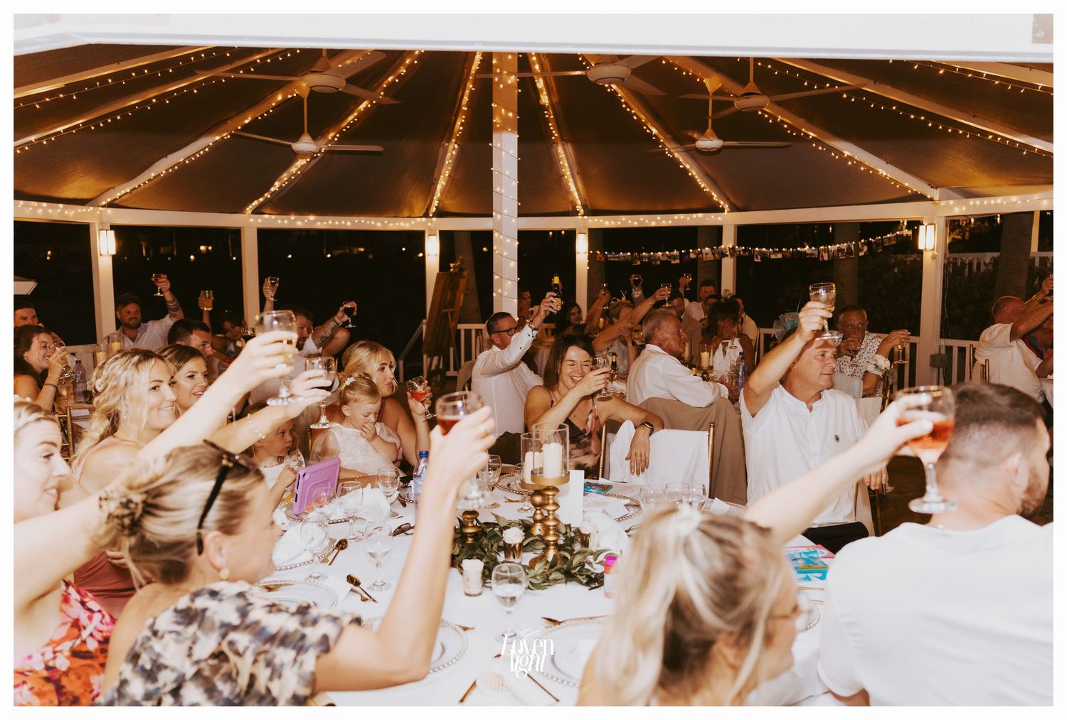 Wedding guests raising their glasses for a toast under a lit tent canopy.