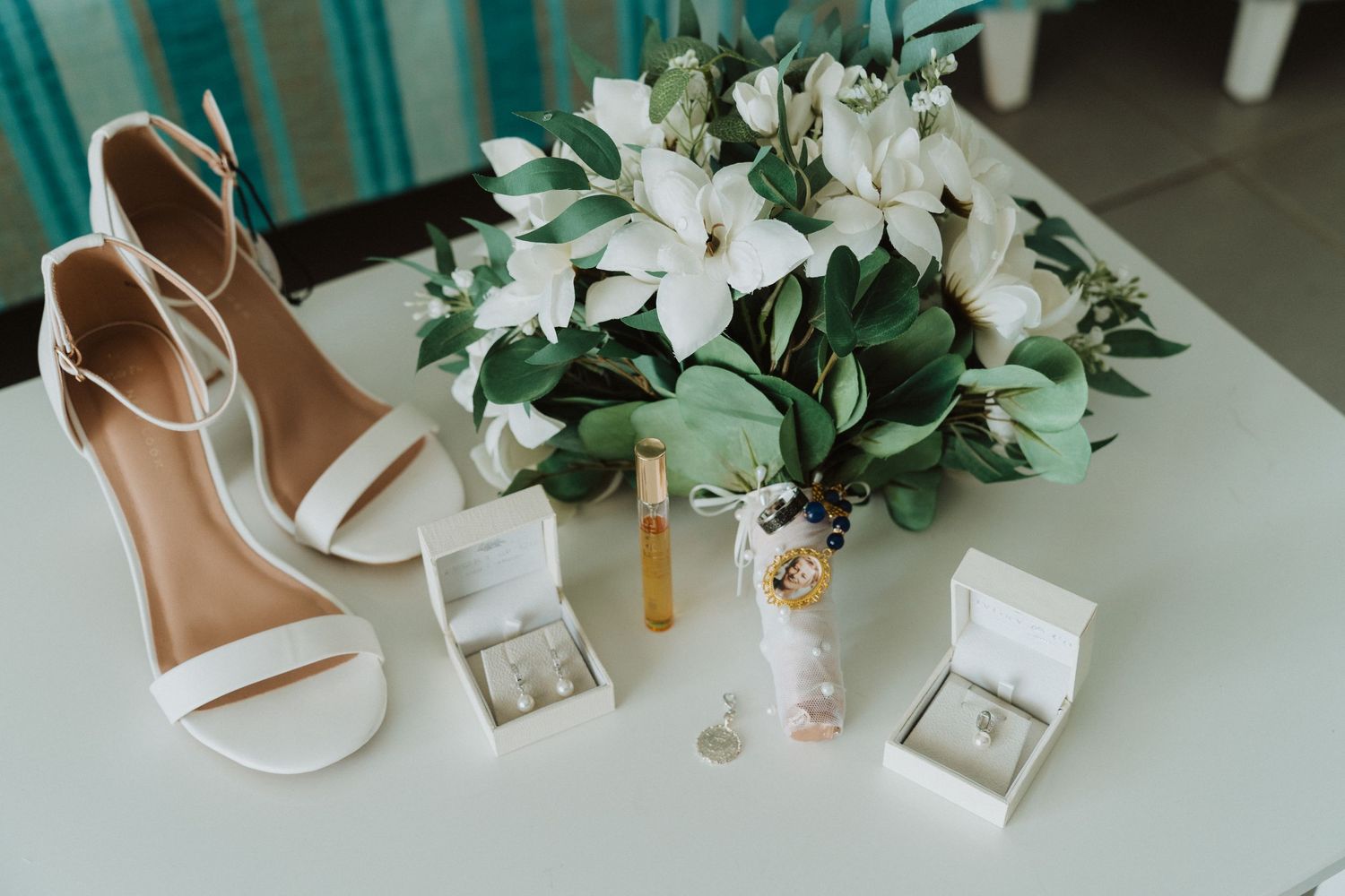 White bridal shoes, jewelry boxes, and white lily floral arrangement on light surface with turquoise background.