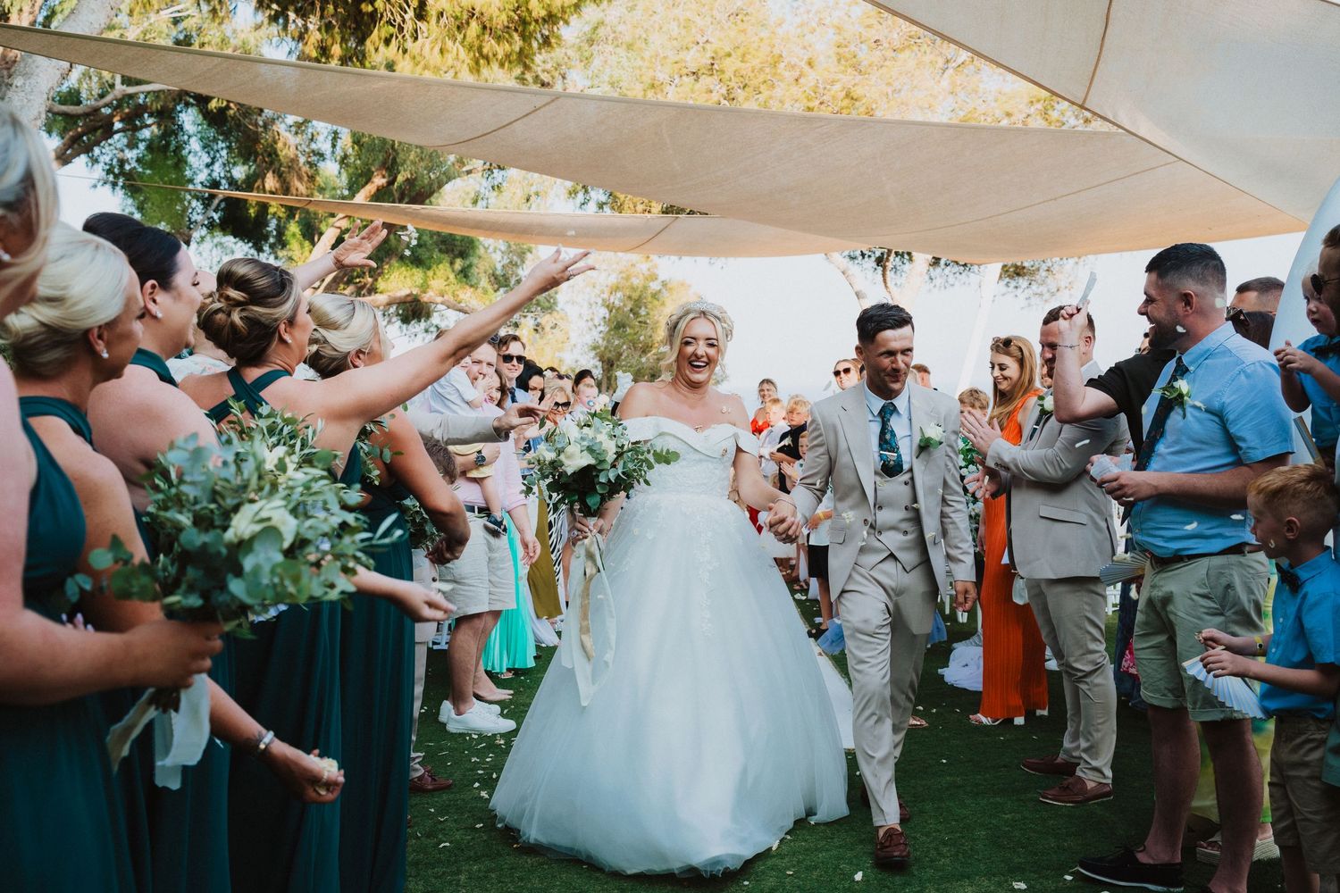 Wedding guests celebrate with bubbles as newlyweds walk down an outdoor aisle under a white canopy during daytime ceremony.