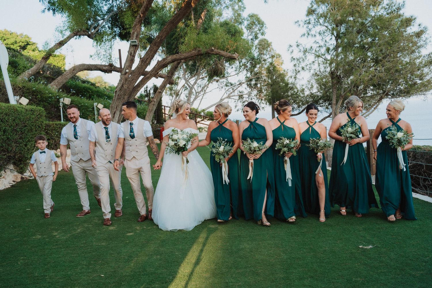 Wedding party in white and emerald green formal attire poses together on a grassy outdoor venue by trees.