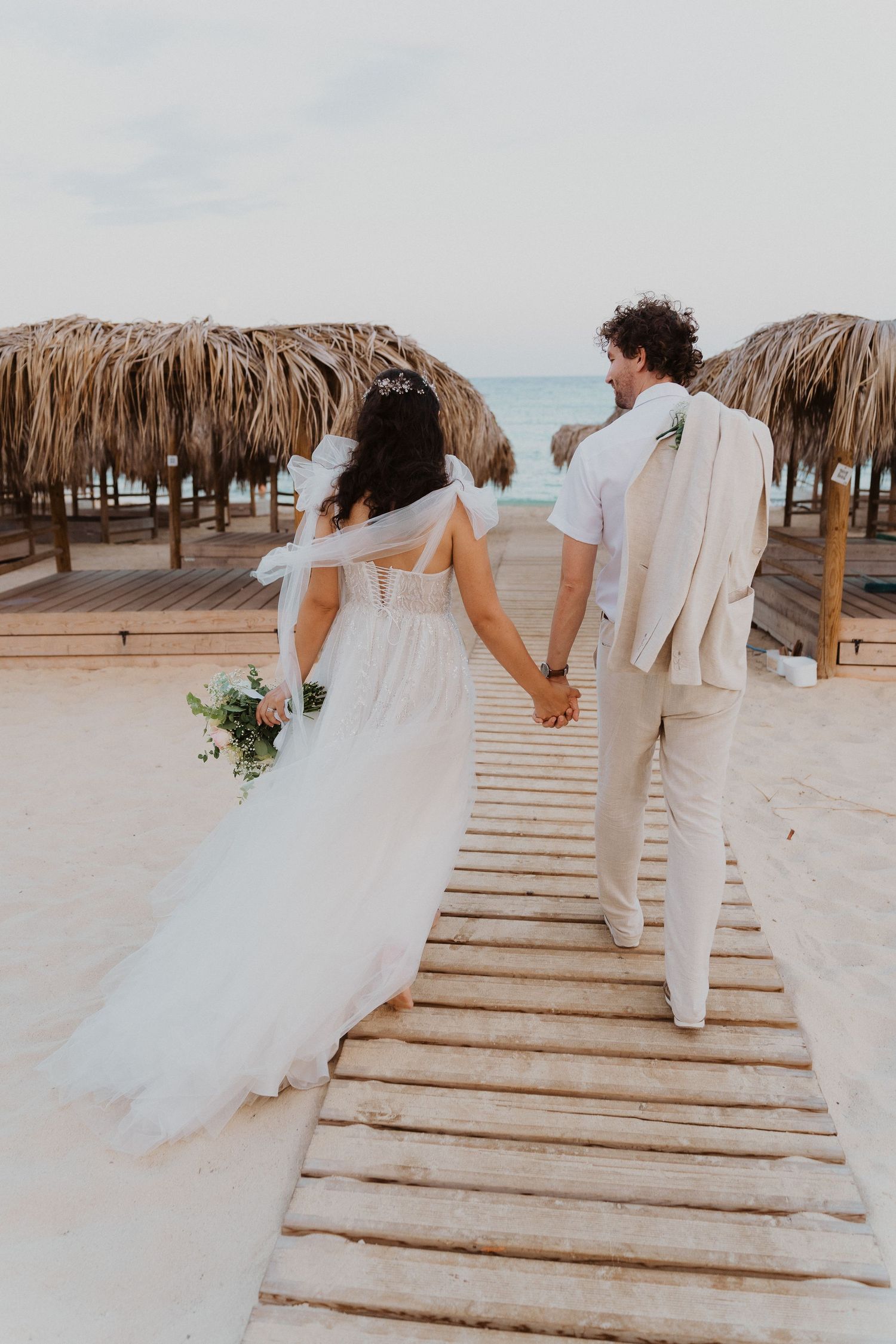 Couple walking on wooden boardwalk between tiki umbrellas at beach wedding.