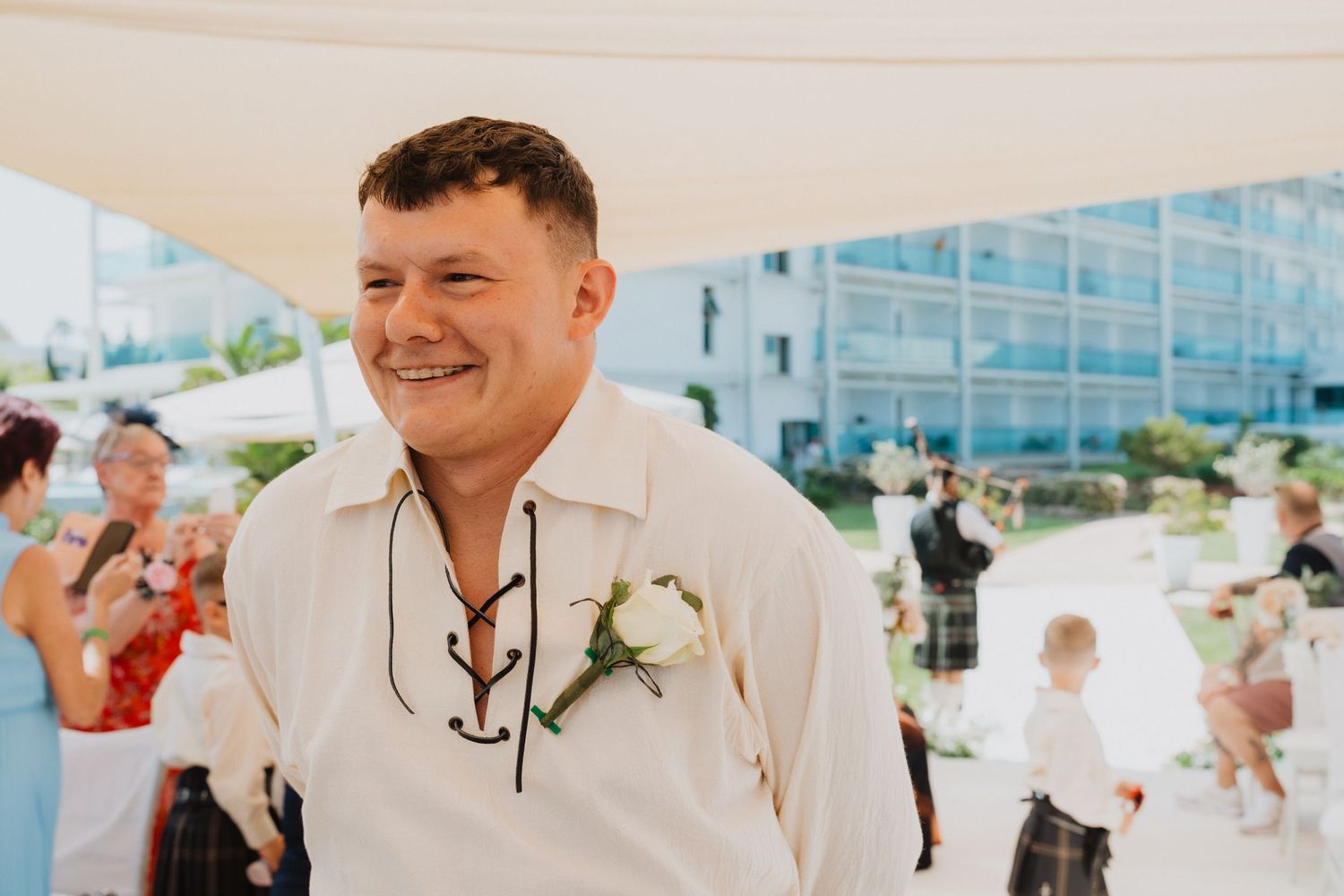 Wedding guest in traditional Scottish attire smiling outdoors during ceremony.