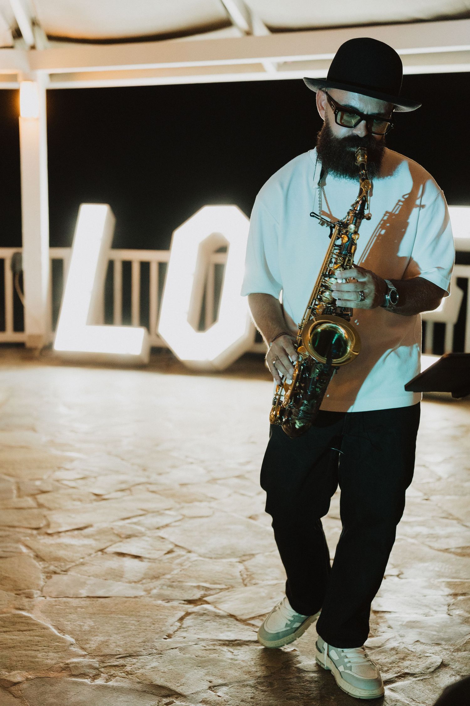 A musician in casual attire and fedora plays saxophone on stage with a glowing LOVE sign in the background.