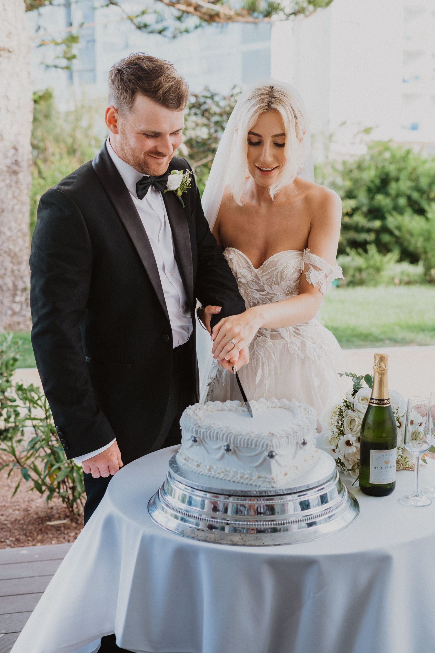 Couple cutting their elegant white wedding cake on covered patio.