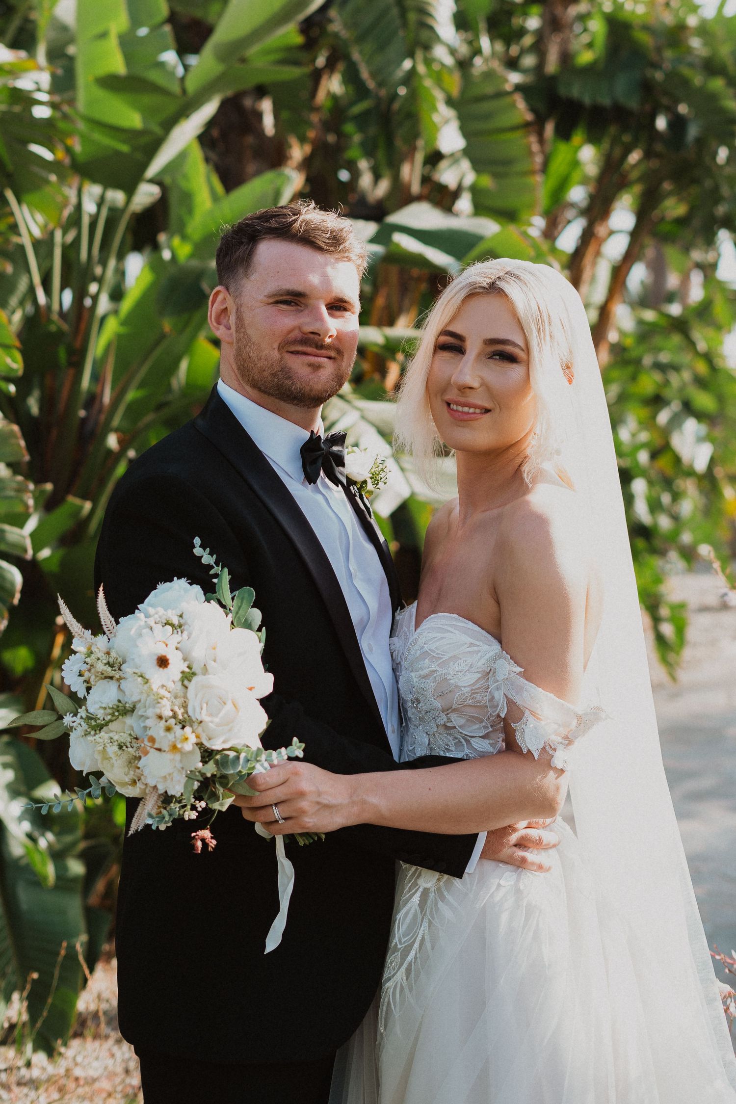 A wedding couple poses outdoors in a tropical garden setting with a white floral bouquet.