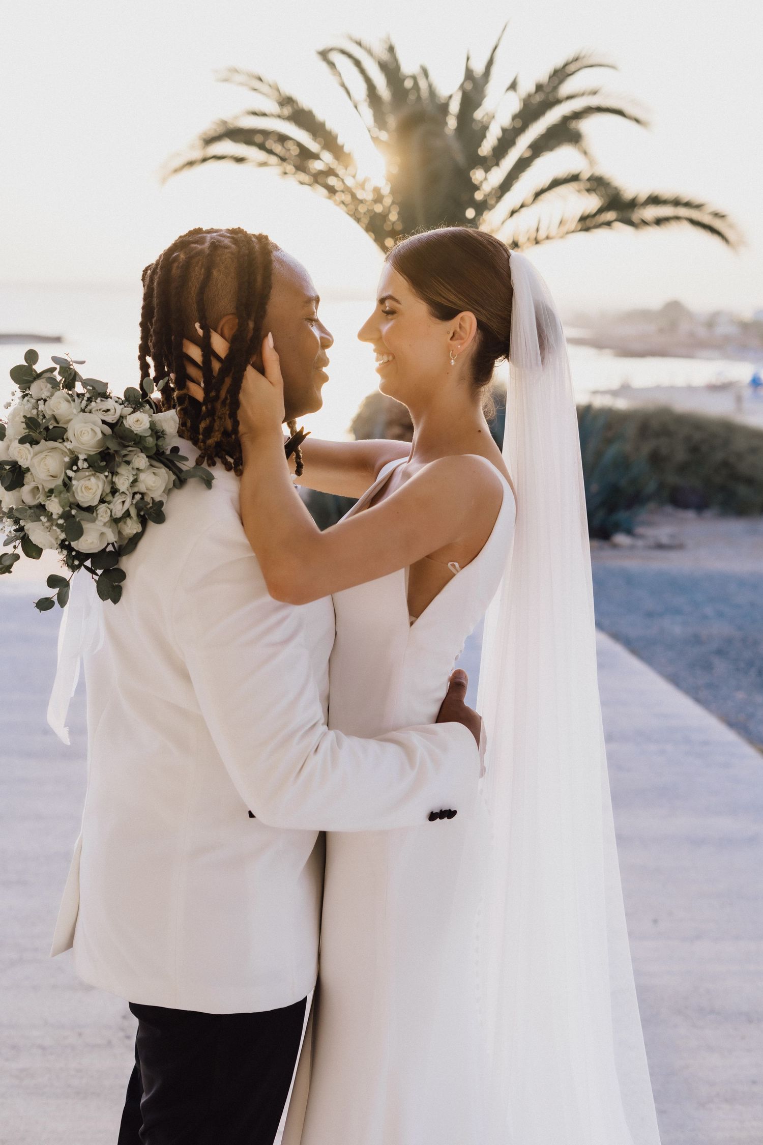 A tender wedding moment captured at sunset with palm trees and bouquet of white flowers.