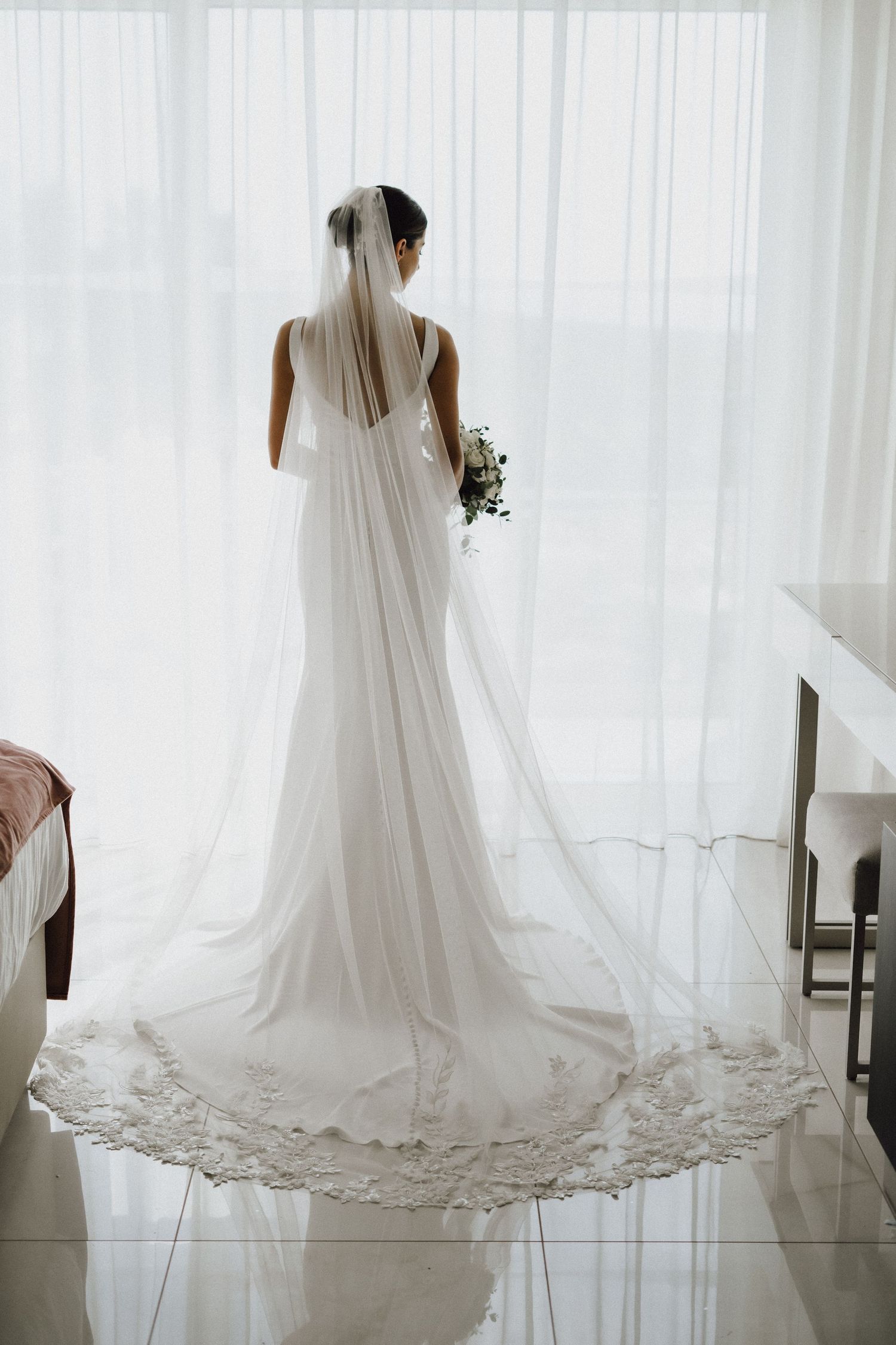 Back view of bride in flowing white wedding gown holding bouquet against sheer curtain backdrop.