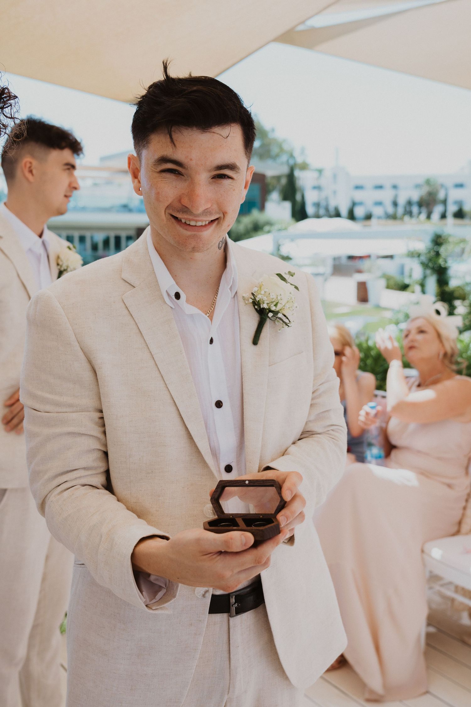 A wedding guest in a beige suit and white shirt holds a ring box while smiling during an outdoor celebration.