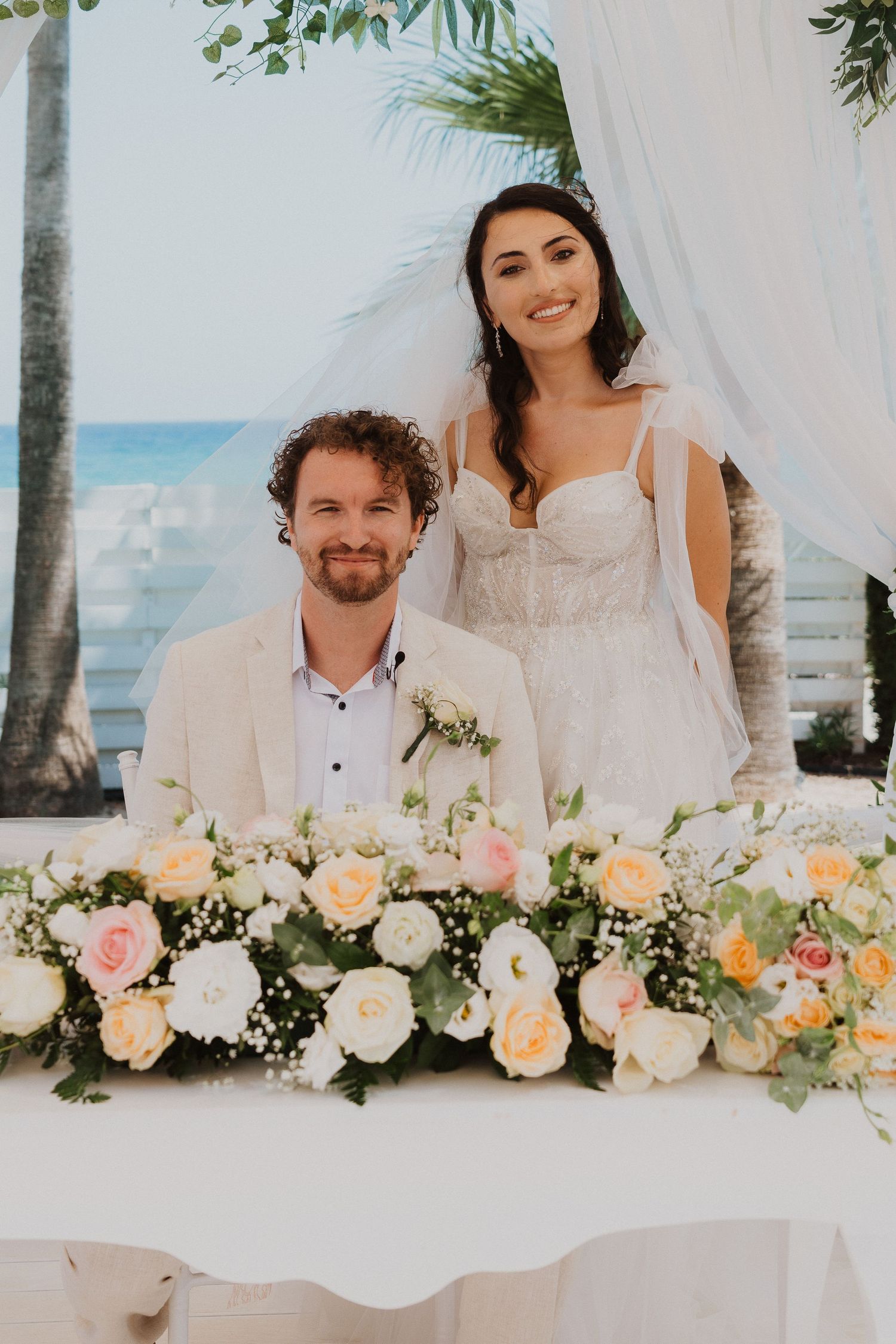 Newlyweds pose together at a beach wedding ceremony with elegant peach and white floral arrangements.