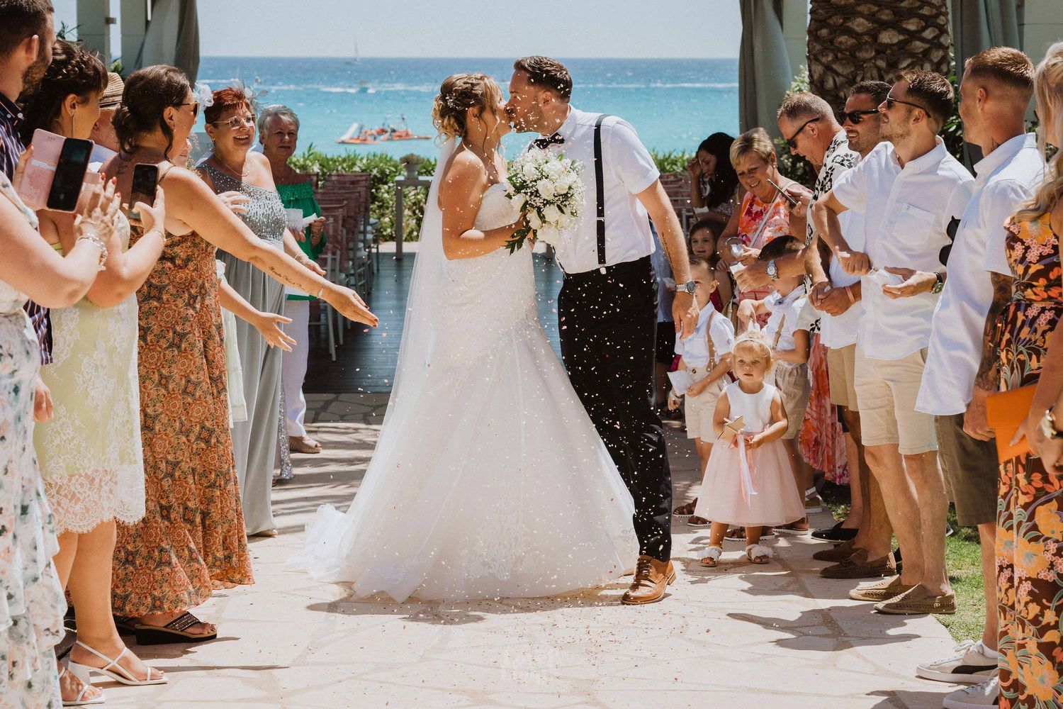 Wedding exit celebration with guests throwing confetti in Mediterranean beachside setting.