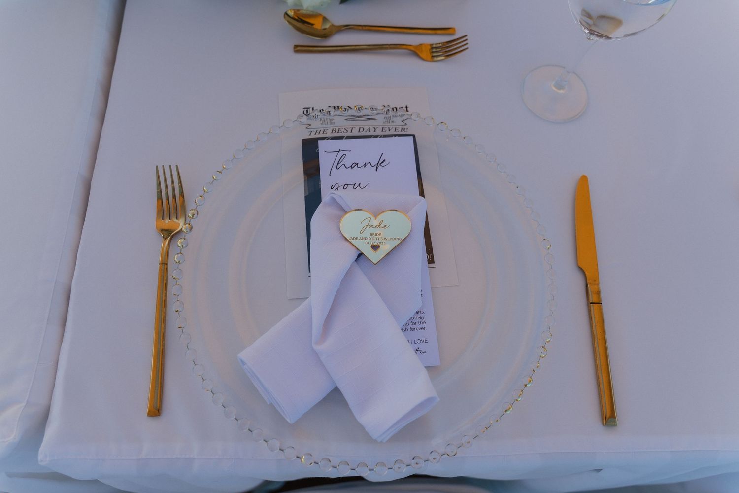 Place settings at a wedding reception feature gold cutlery and white napkins arranged on a white tablecloth.
