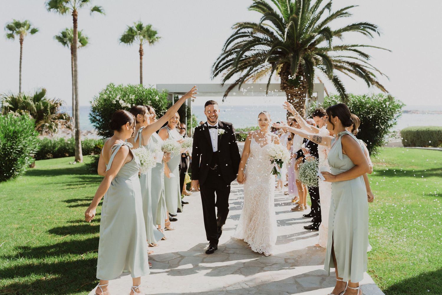 Wedding party walking in formation on a manicured lawn with palm trees and elegant decor.