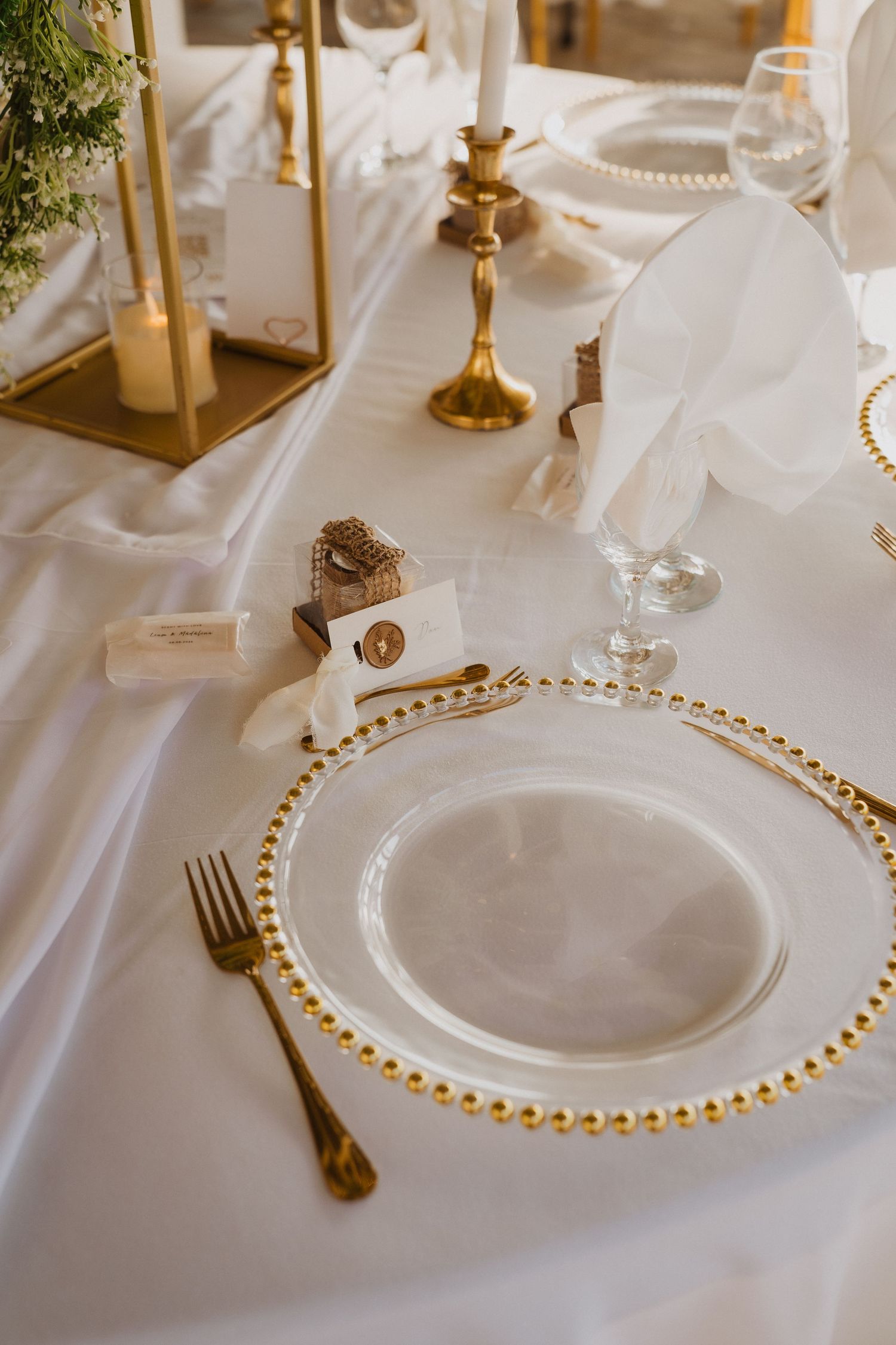 Elegant wedding table setting with gold beaded charger plates, gold cutlery, white napkins, and brass candlesticks on white linen.