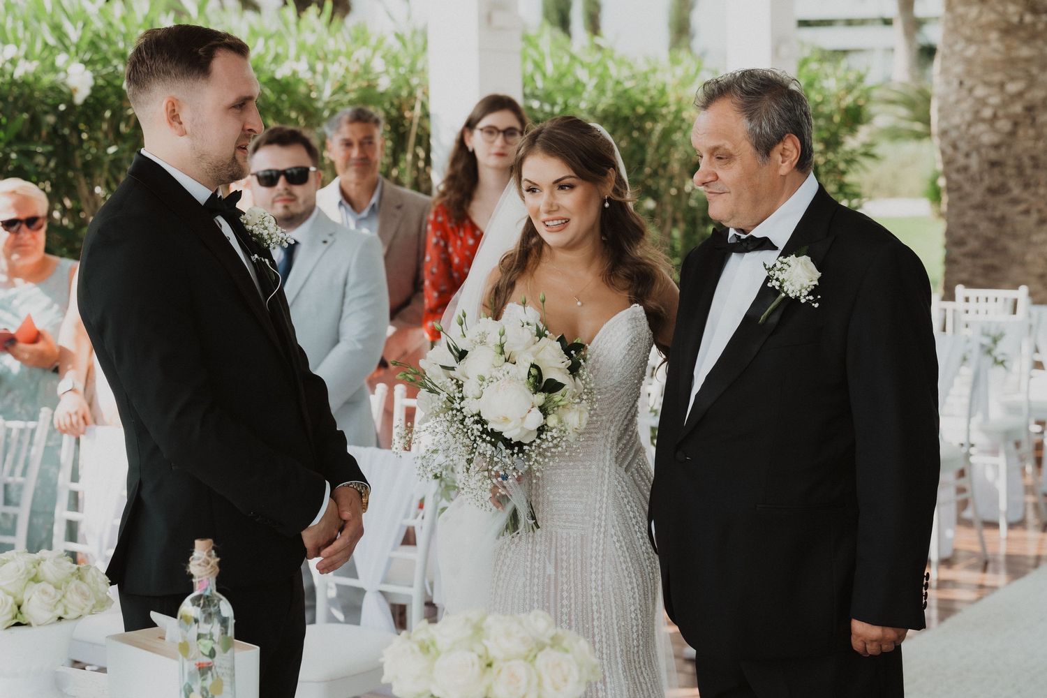 Bride in beaded gown meeting groom at outdoor wedding ceremony altar with father.