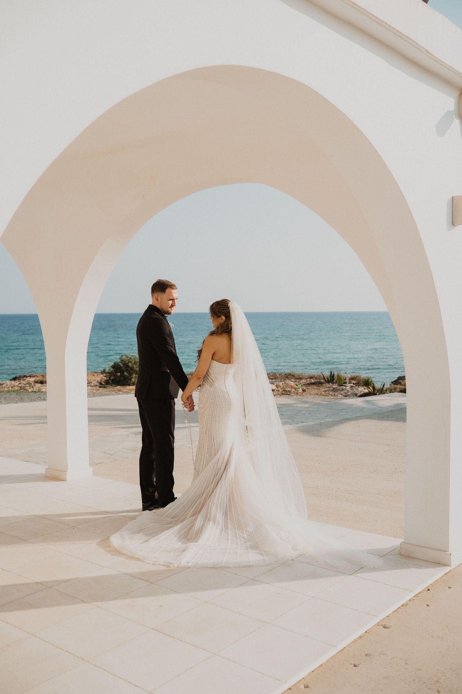 Wedding couple holding hands under a white arch overlooking the turquoise Mediterranean sea.