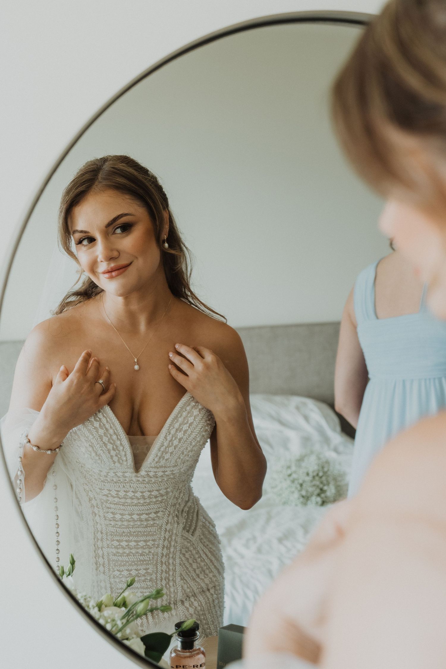 Bride in beaded gown adjusting necklace while gazing at her reflection in a round mirror.