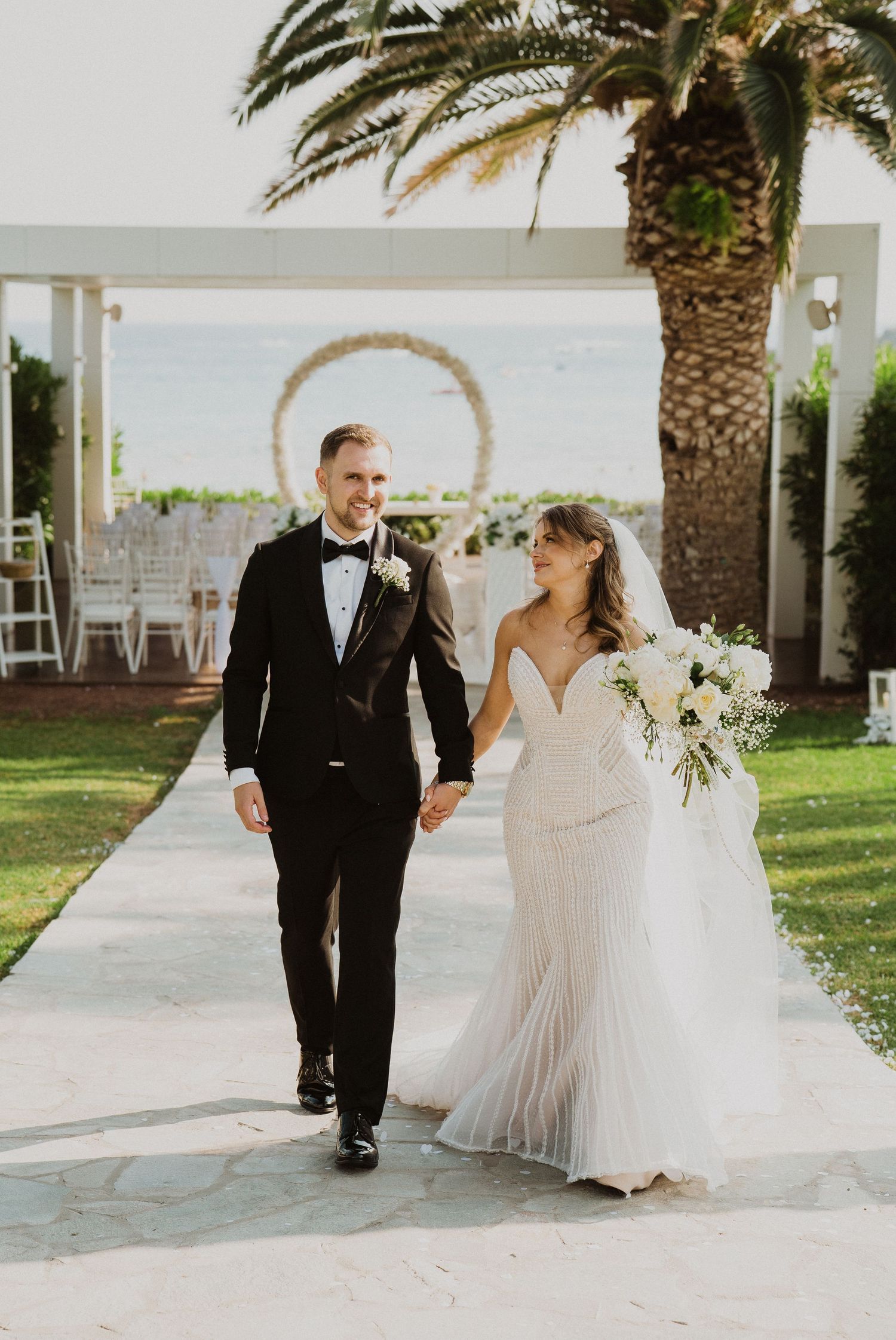 Bride and groom walk hand-in-hand at outdoor wedding ceremony with circular floral arch and palm trees by the sea.