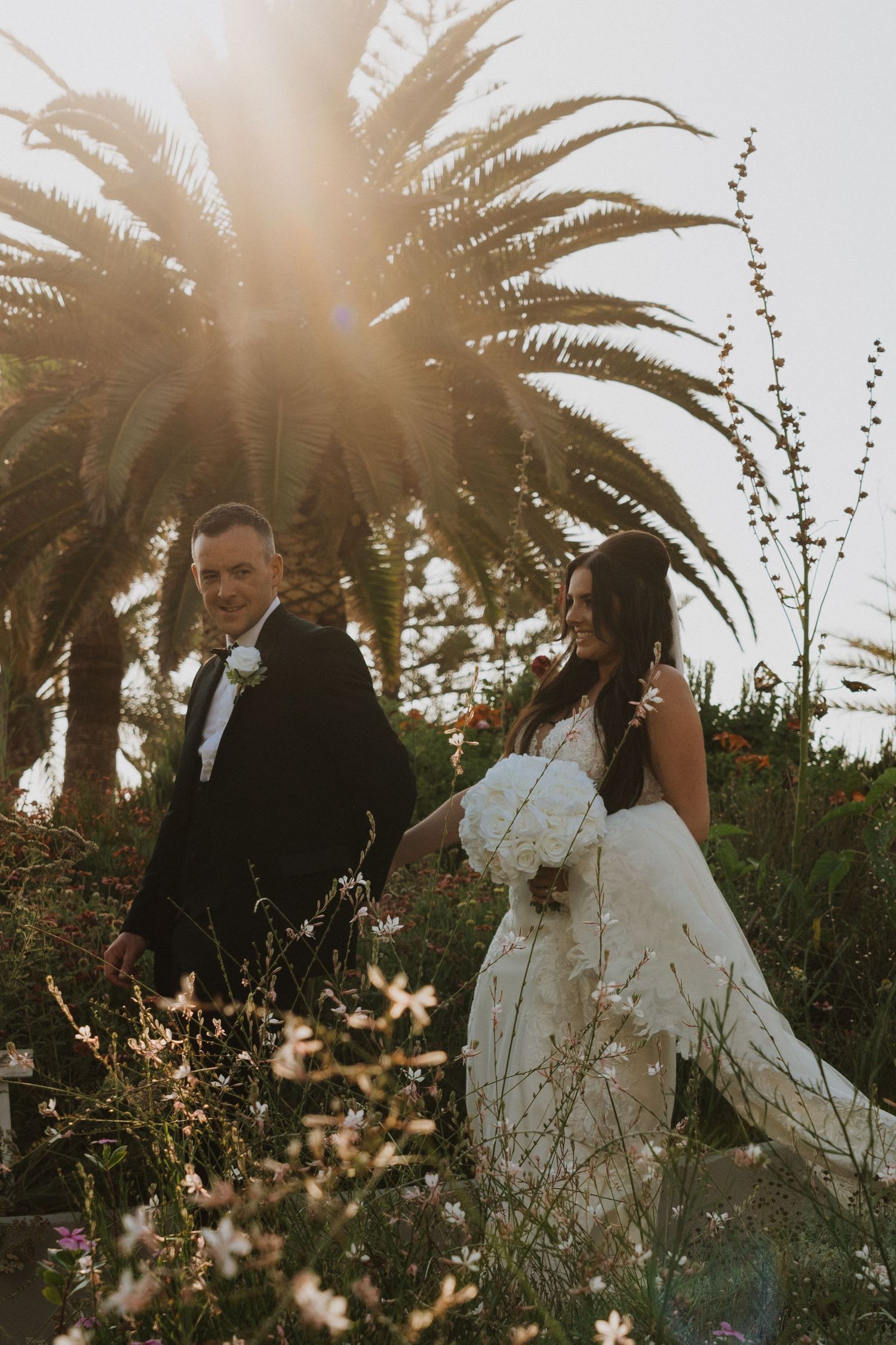 A wedding couple walks through a sunlit garden with towering palm trees and wildflowers.