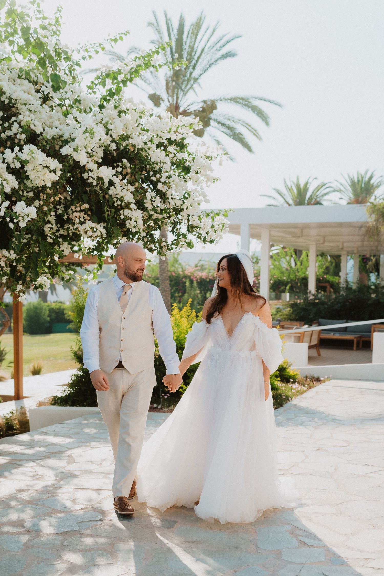 A couple walks together on a sunny stone pathway surrounded by palm trees and white flowers.