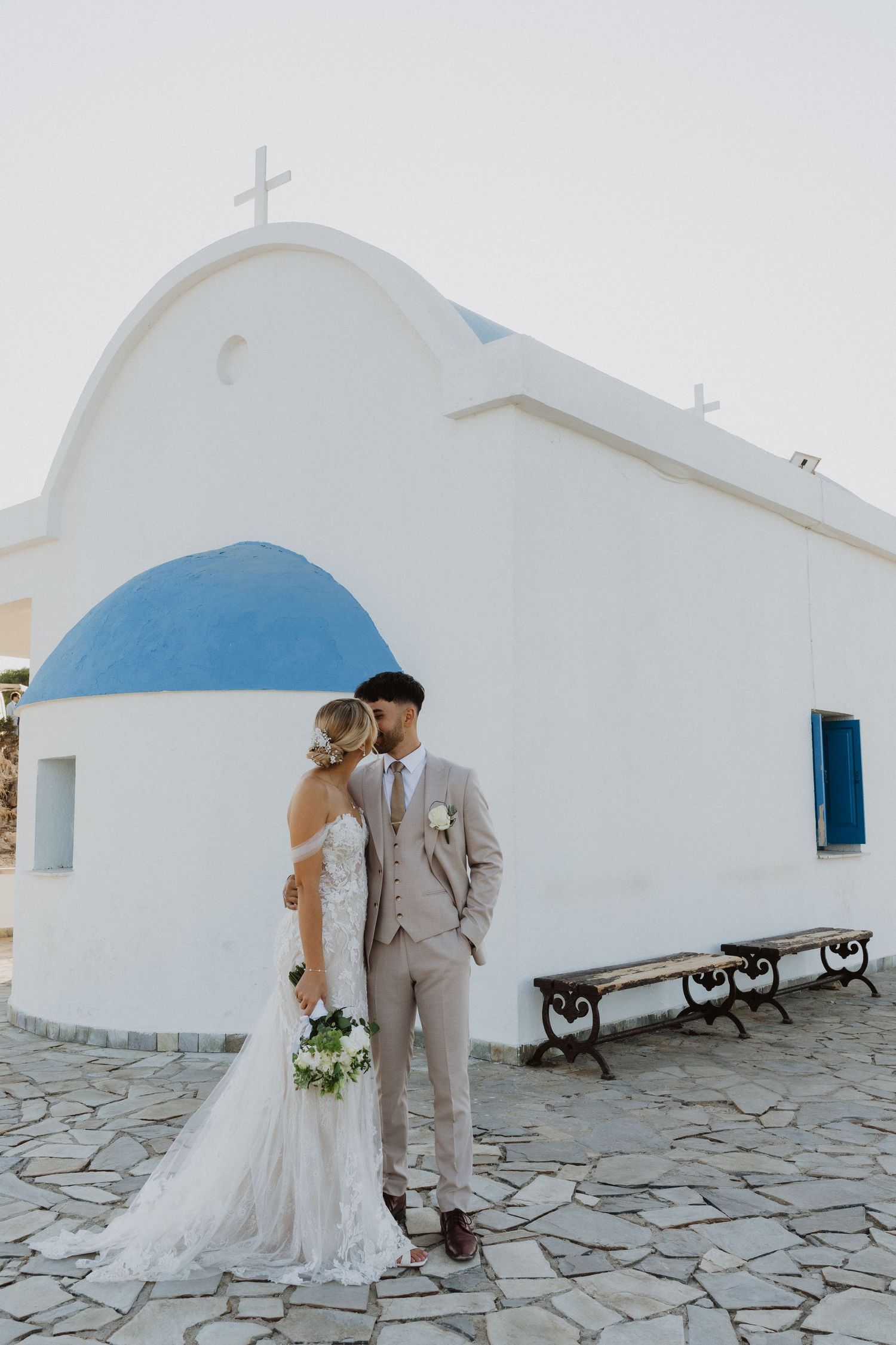 Couple poses by traditional Greek orthodox church with white walls and blue dome.