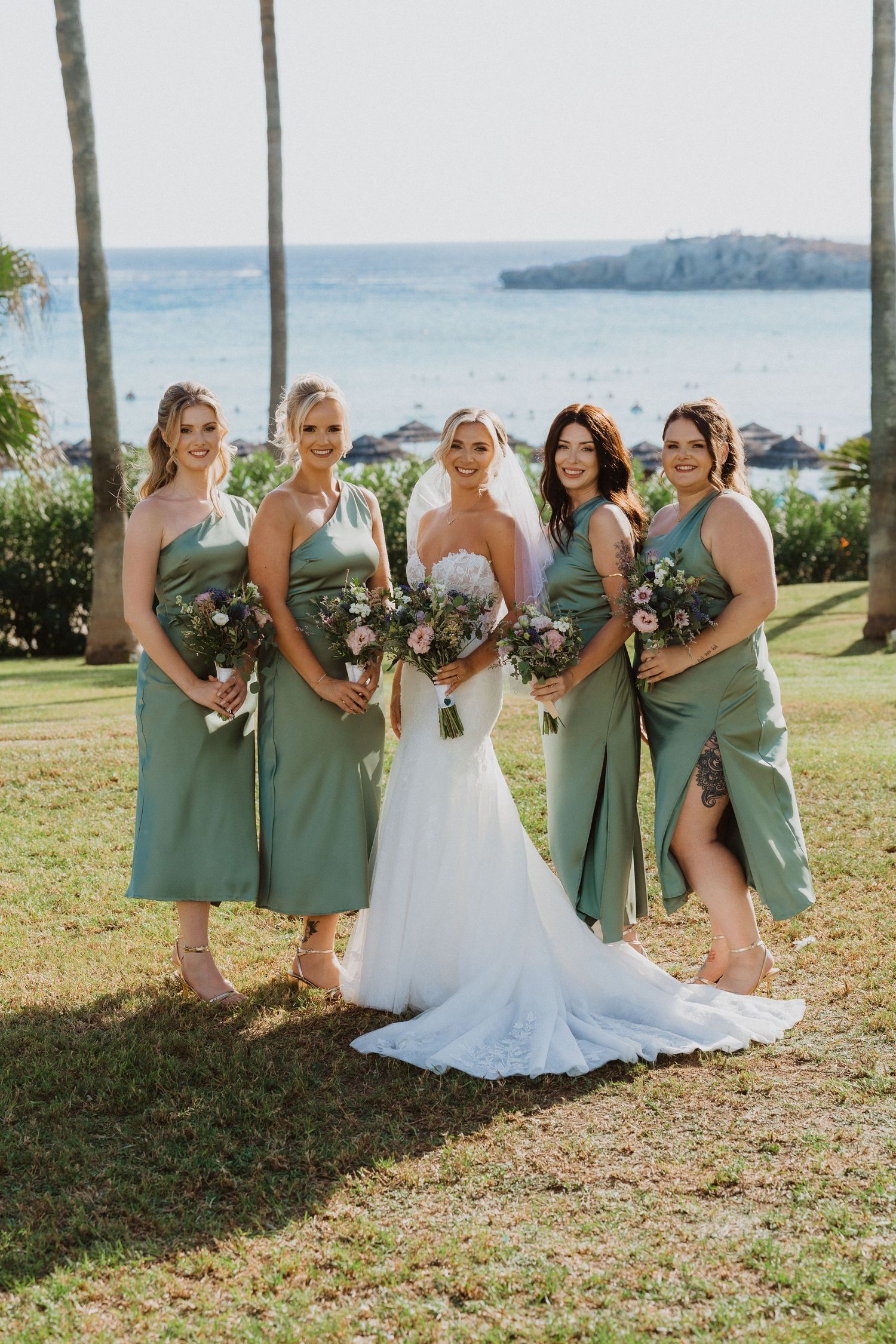 Bridal party in sage green dresses posing together on a grassy oceanfront setting.