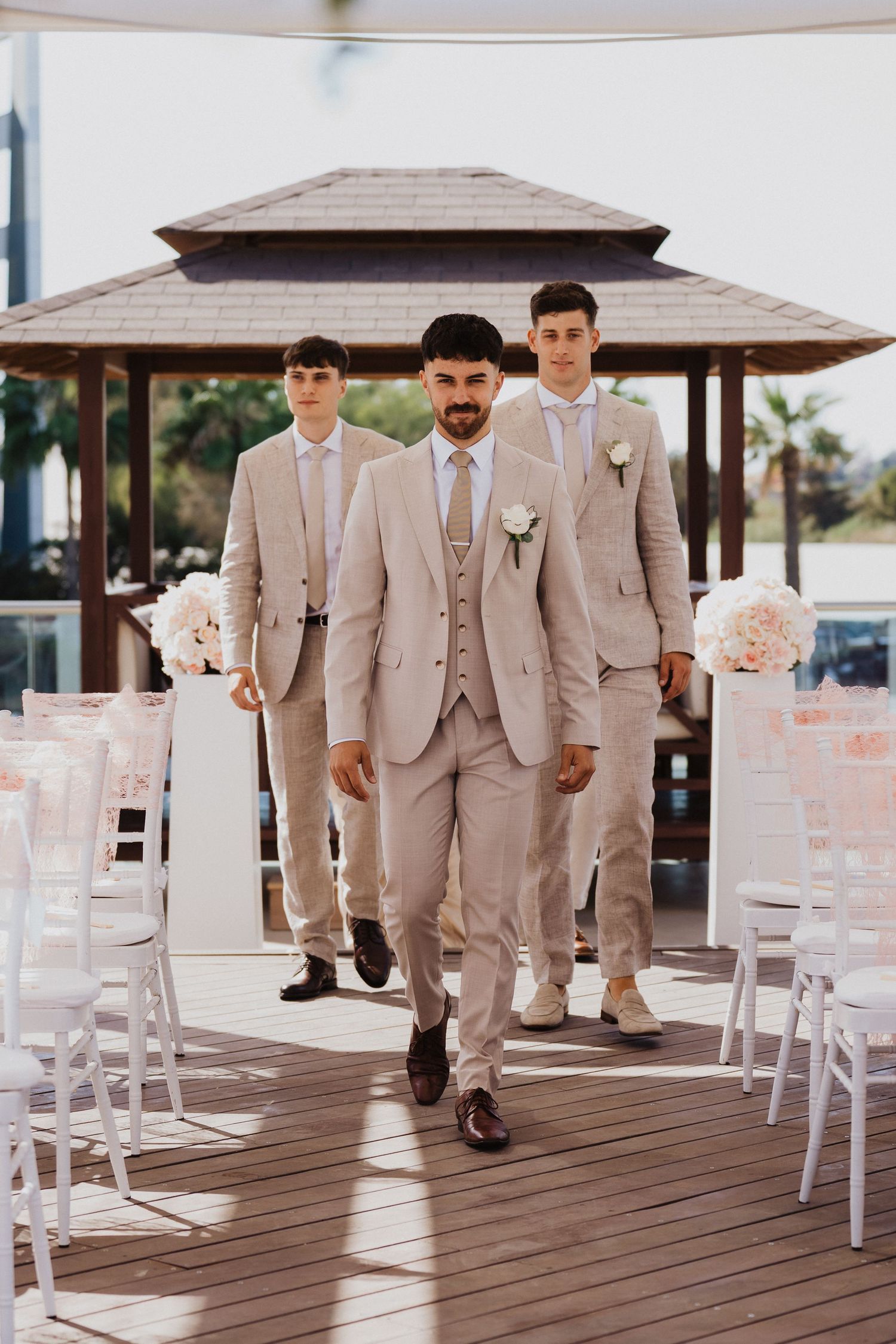 Groomsmen in beige suits walk across a wooden deck with white chairs set up for ceremony.