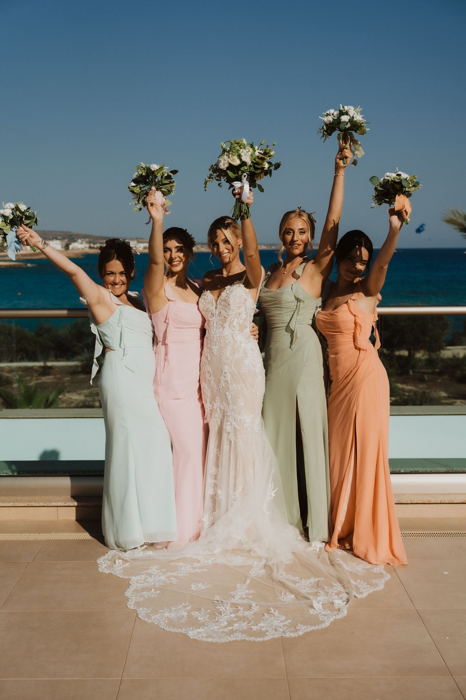 Bridesmaids in pastel gowns raise bouquets while celebrating on a seaside terrace.