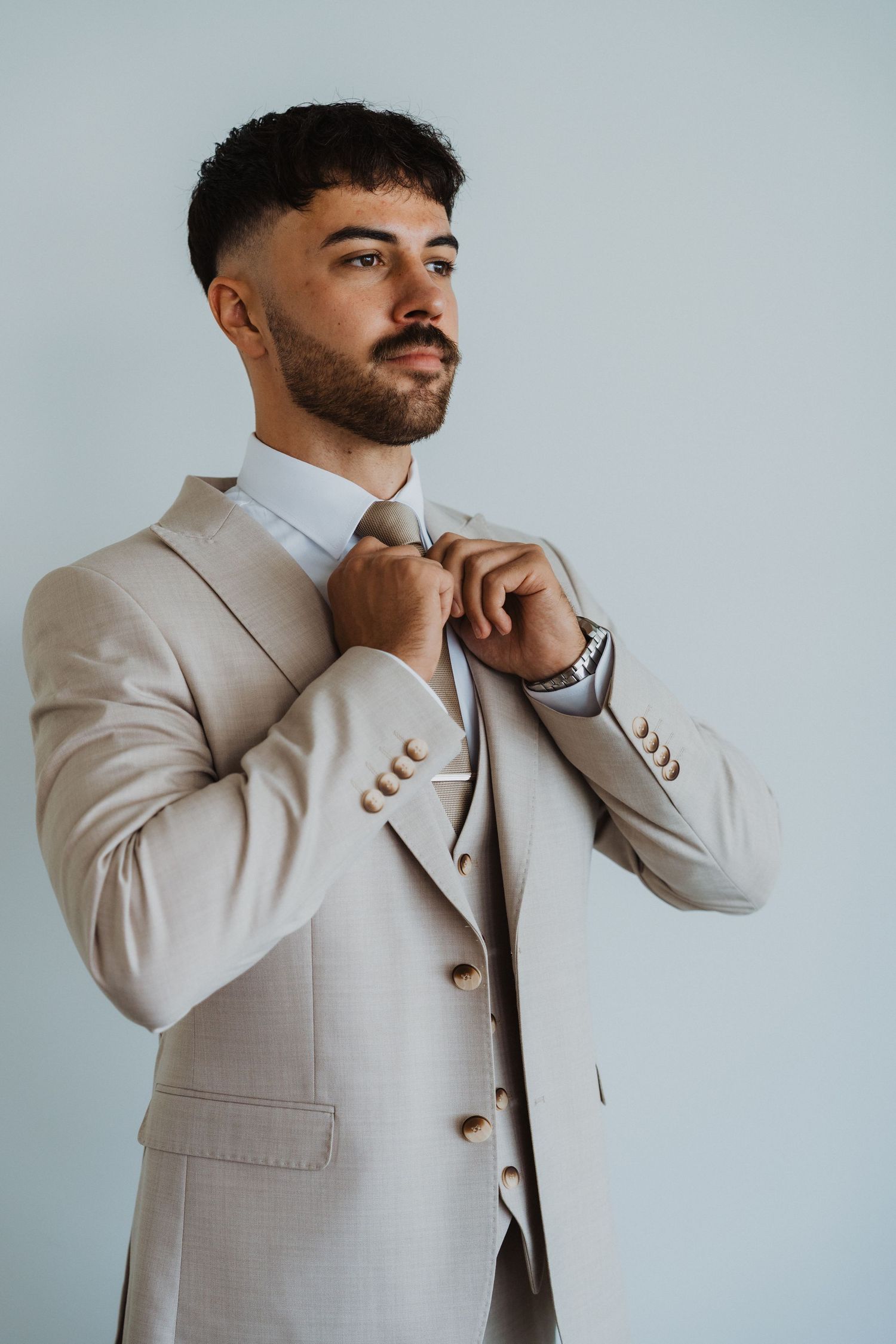 Stylish groom in a light beige suit adjusting his collar against white backdrop.