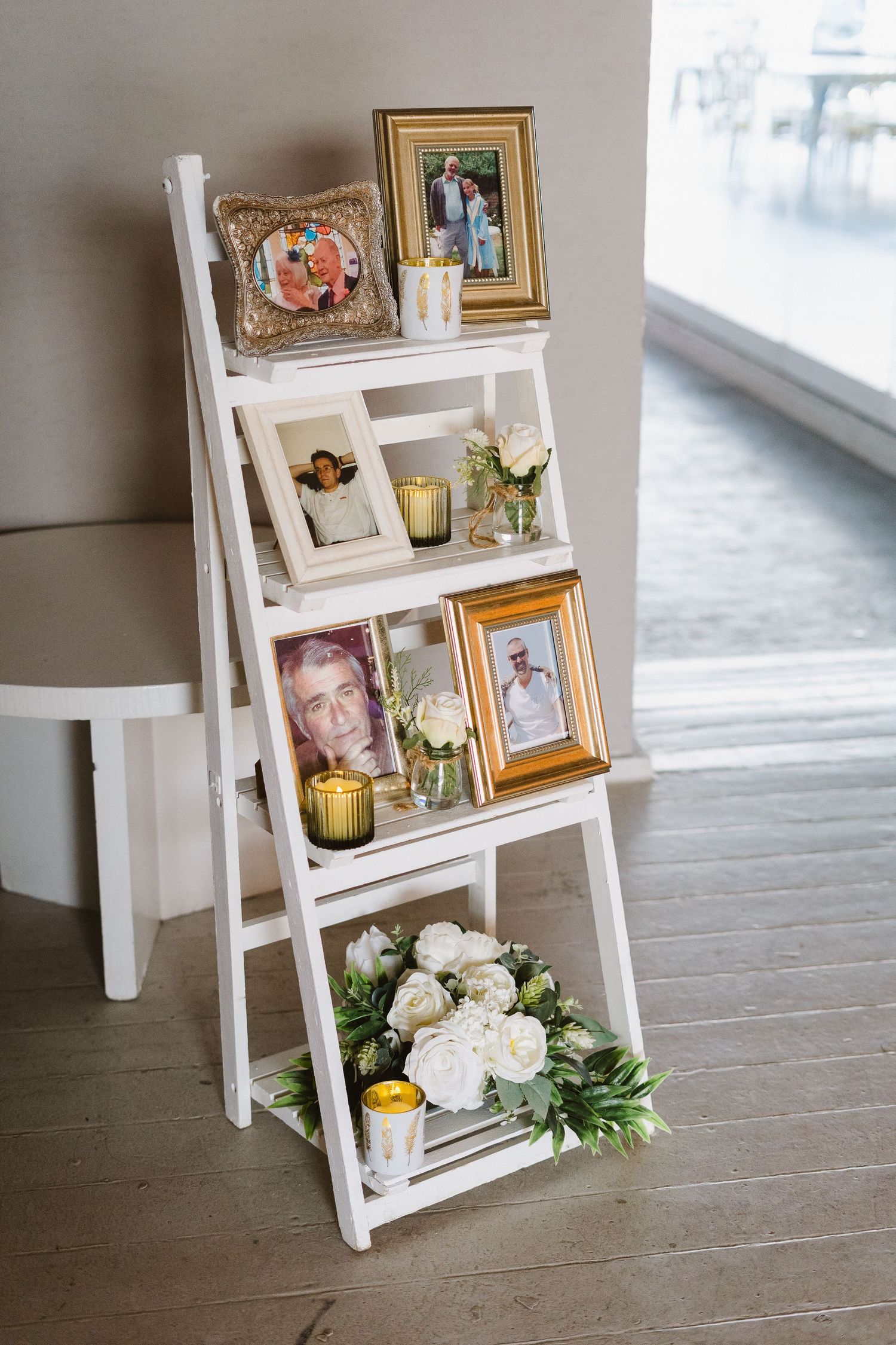 White ladder shelf decorated with framed photos, candles, and white roses for wedding display.