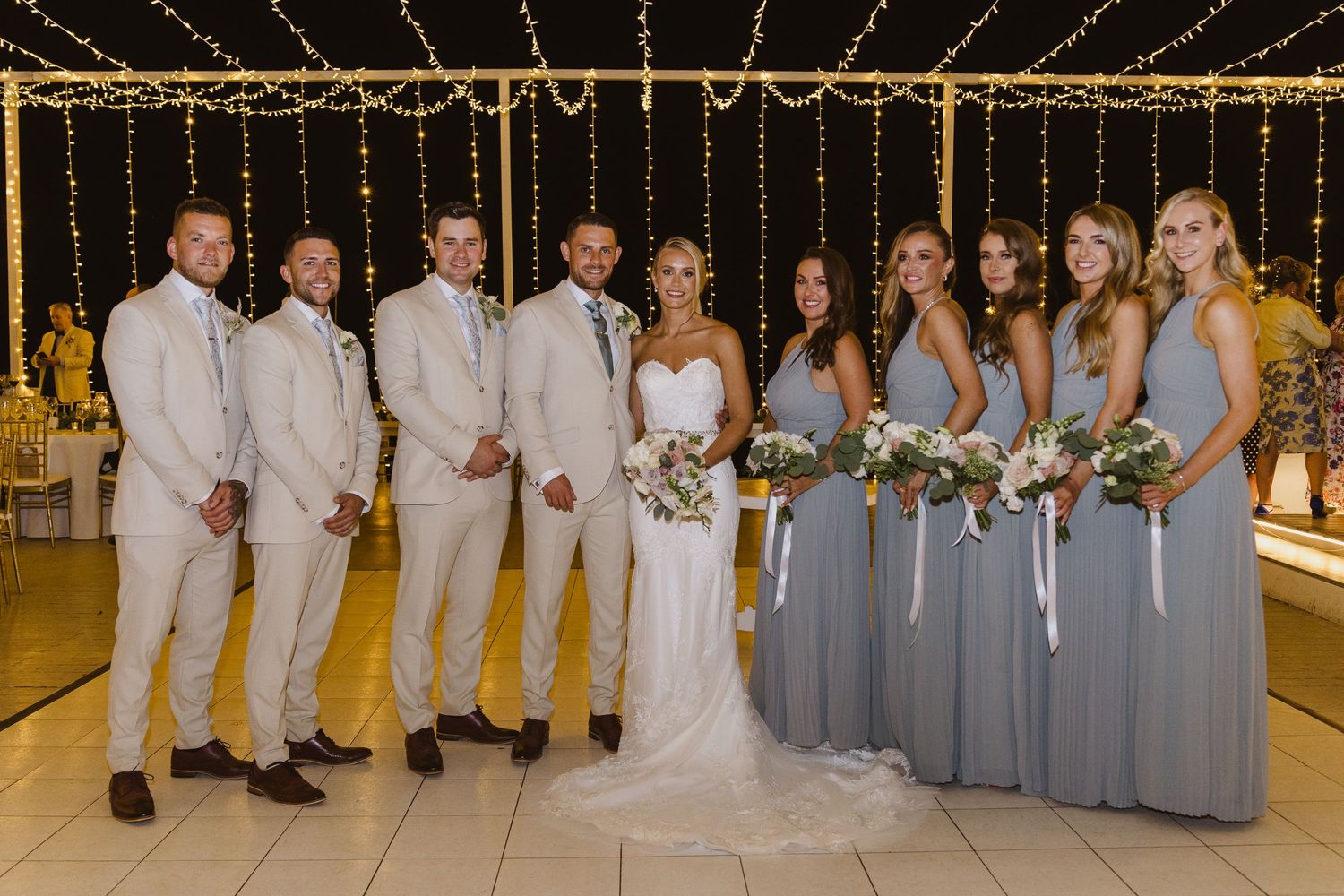 Wedding party in light beige suits and blue-grey dresses poses under string lights at reception.