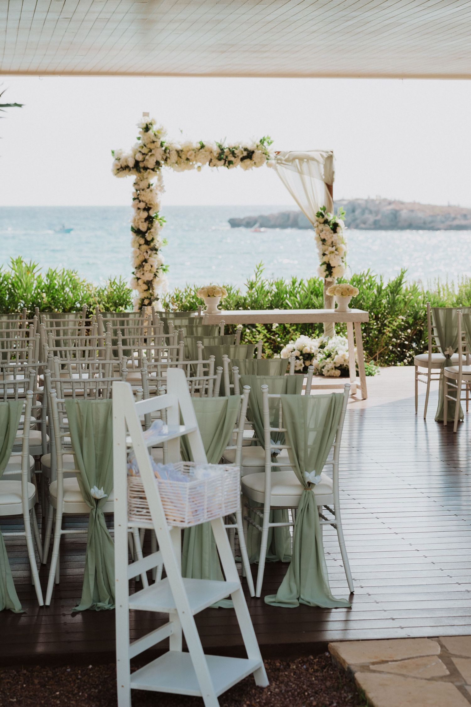 Seaside wedding ceremony setup with floral arch and sage green chair decorations overlooking the ocean.