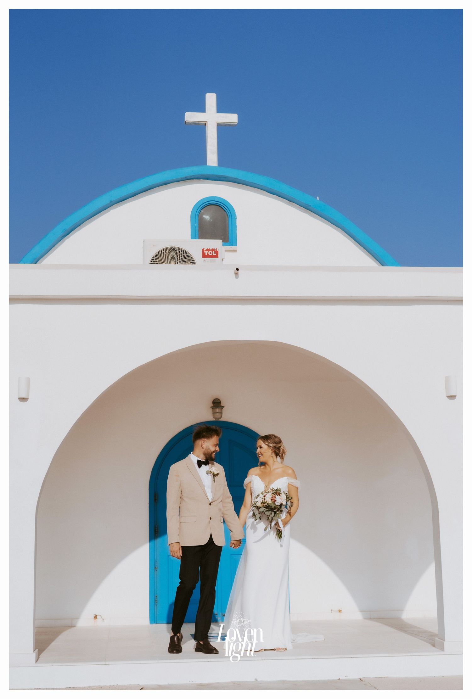 Wedding couple standing in front of traditional Greek chapel with blue door and white architecture.