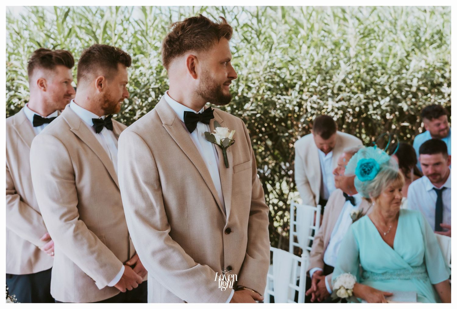 Wedding party stands at outdoor ceremony with groomsmen in beige suits and guest in turquoise.