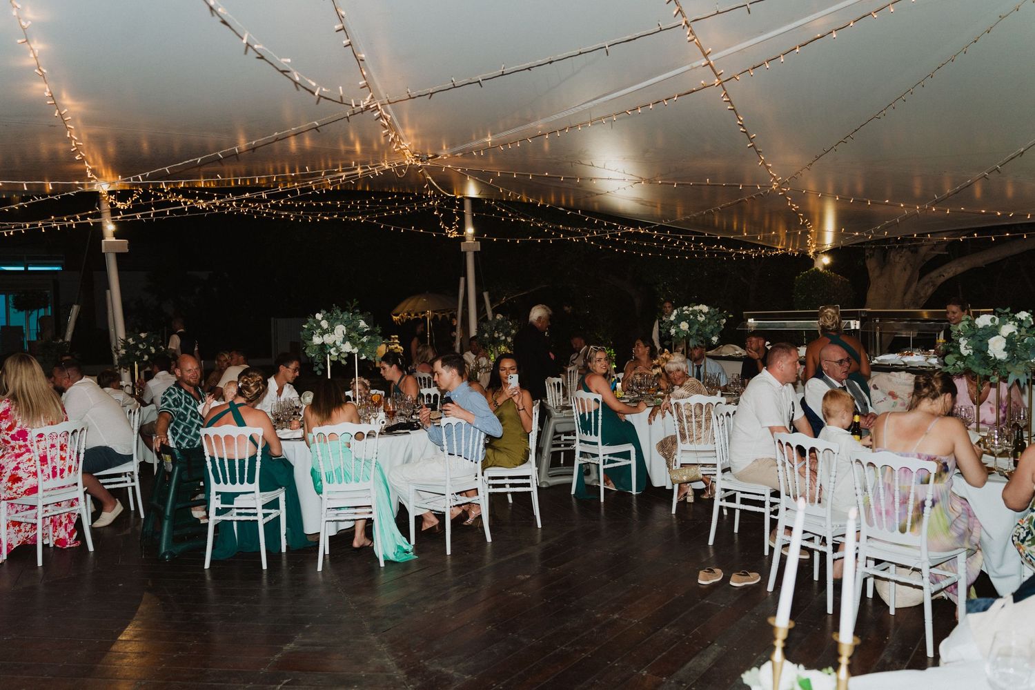 Wedding reception under a tent with twinkling string lights and white chairs arranged around tables.