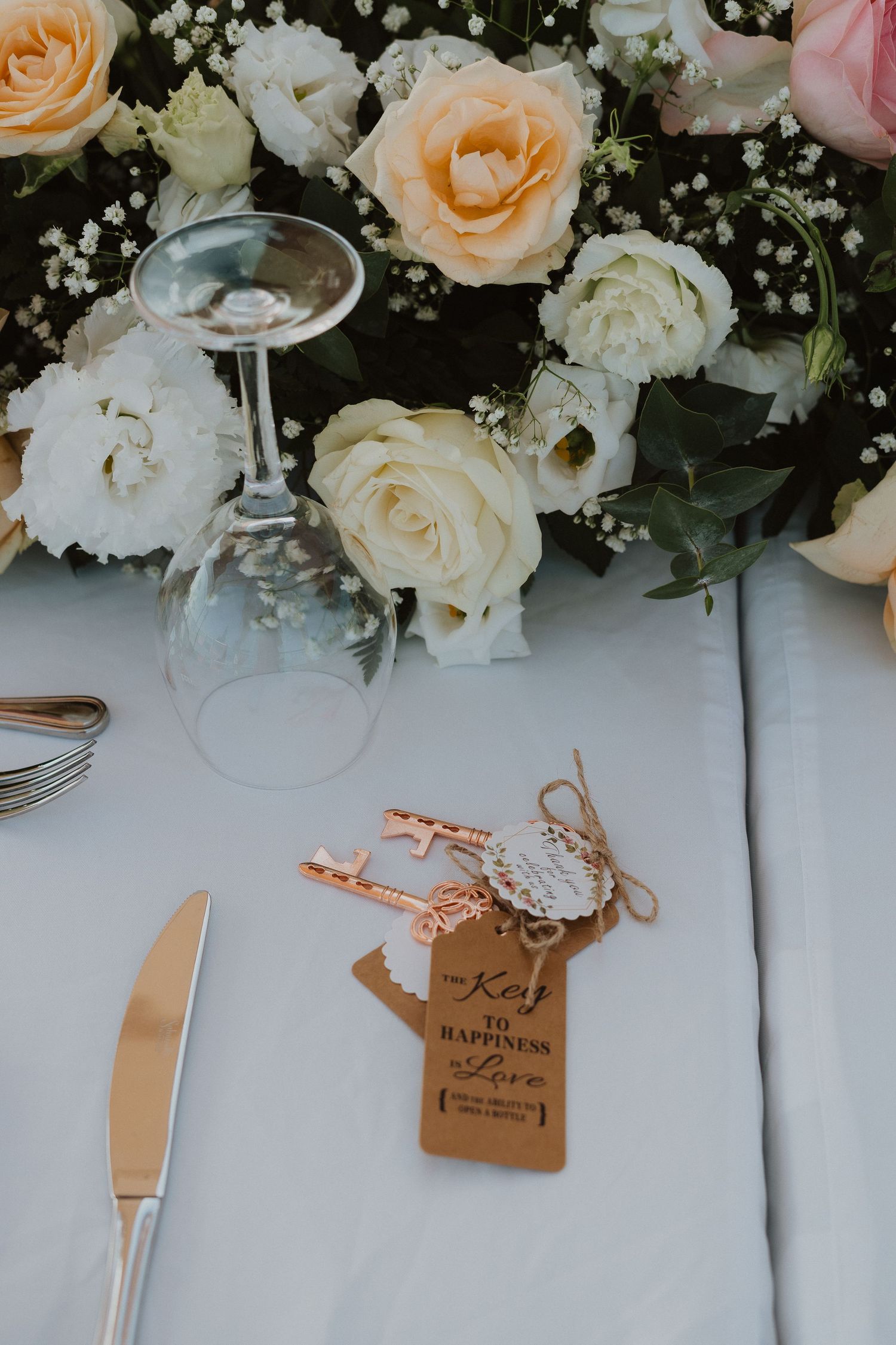 Place setting with rose gold key favor among white and peach roses with wine glass and silverware.