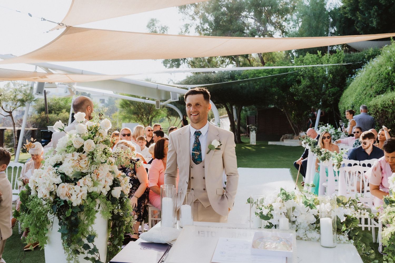 An elegant outdoor wedding ceremony setup with white floral arrangements and cream-colored sail shades overhead.