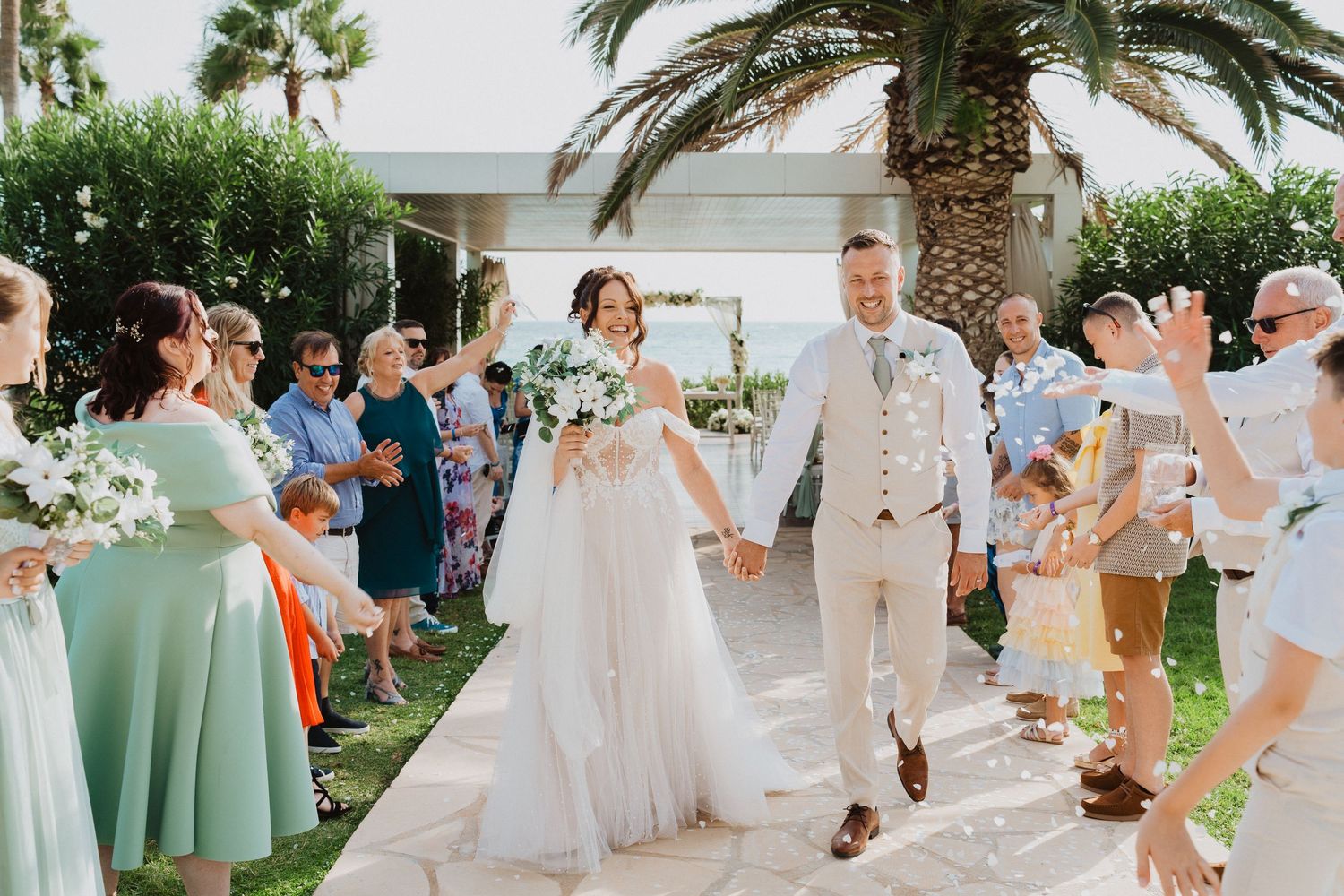 A happy couple walks down a palm-lined outdoor aisle while wedding guests celebrate at a beachside ceremony.