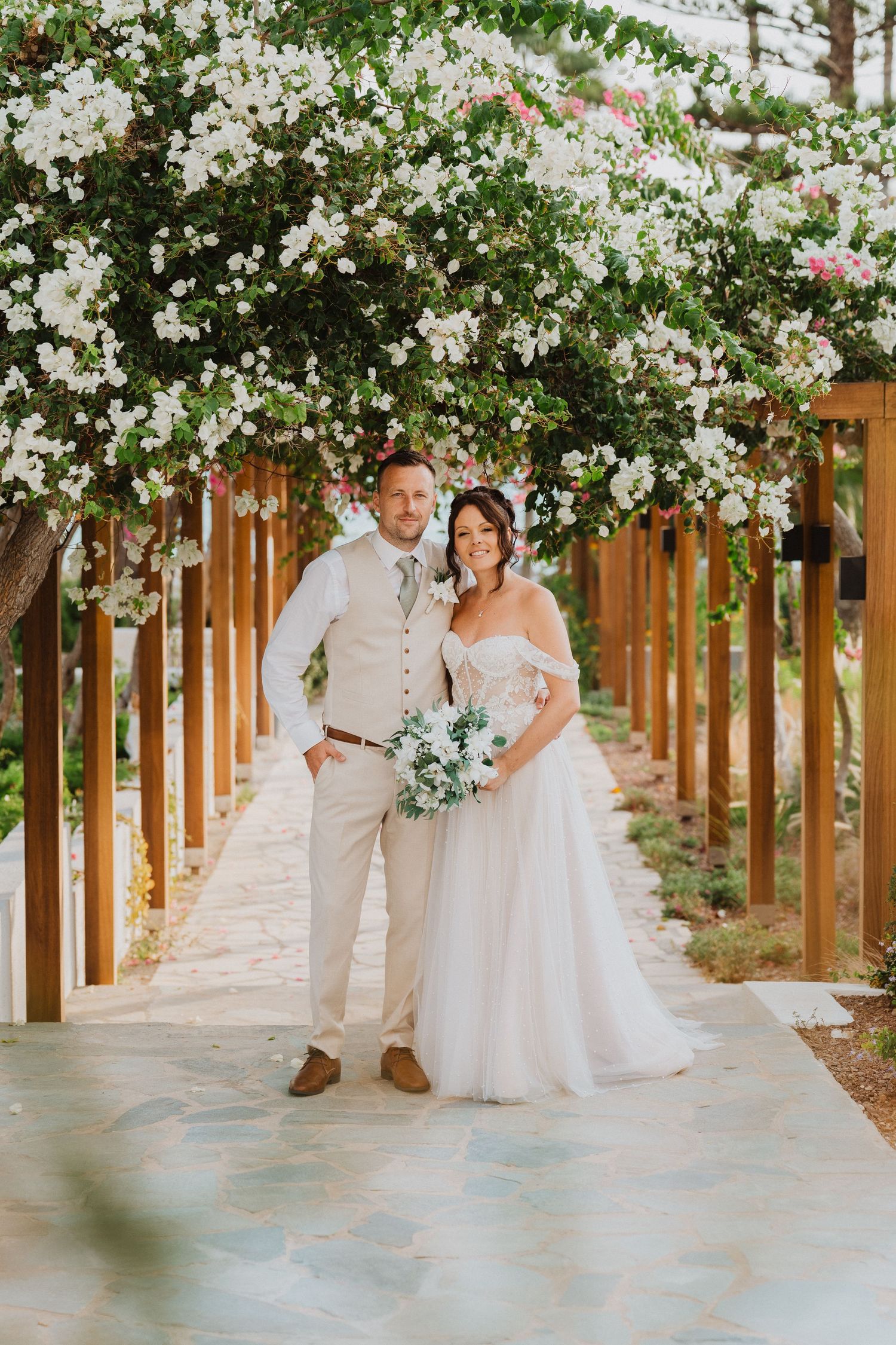 A romantic wedding photo under a blooming white floral arbor with wooden posts on a sunny day.