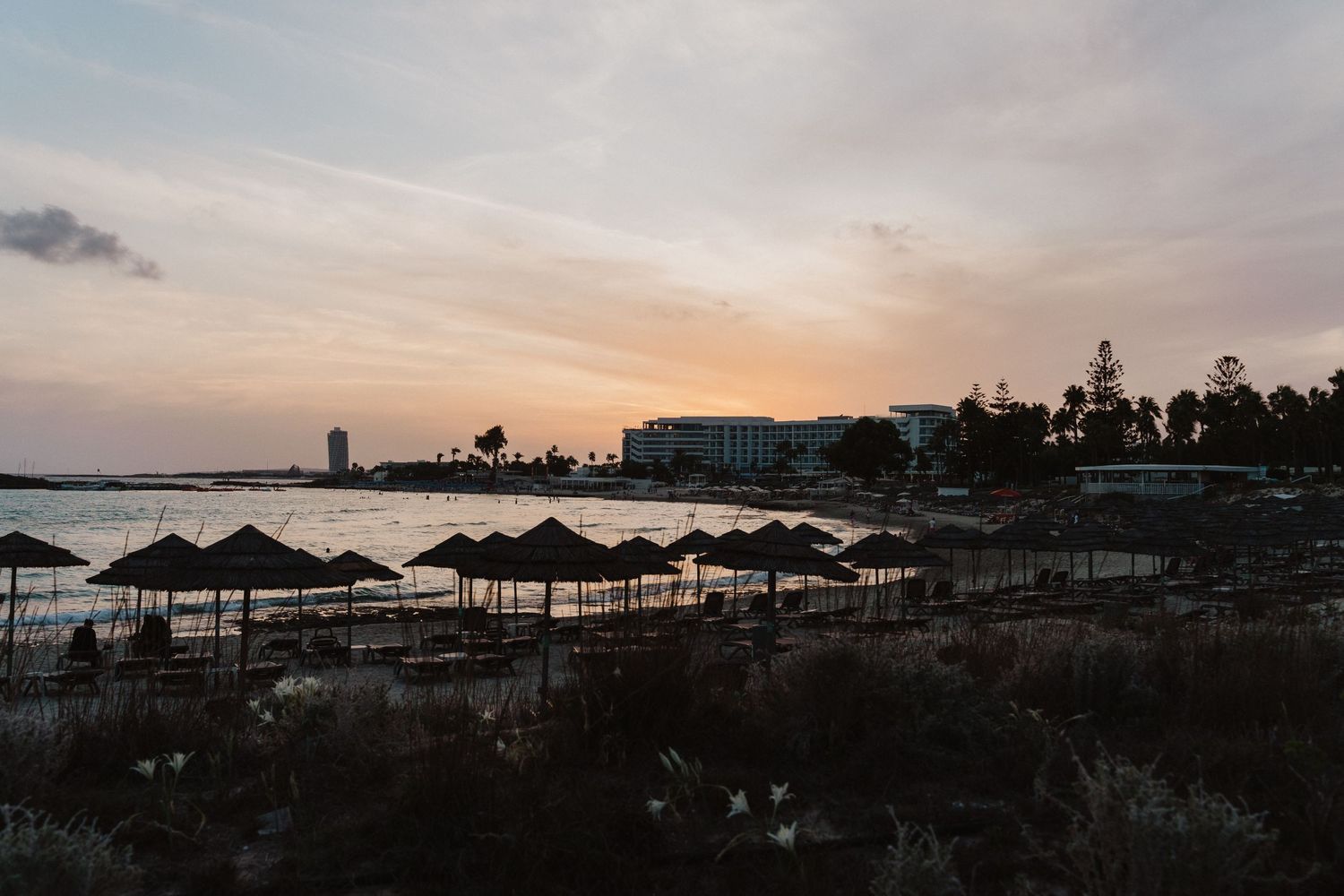 Beach resort silhouetted at sunset with rows of thatched umbrellas and palm trees along the coastline.