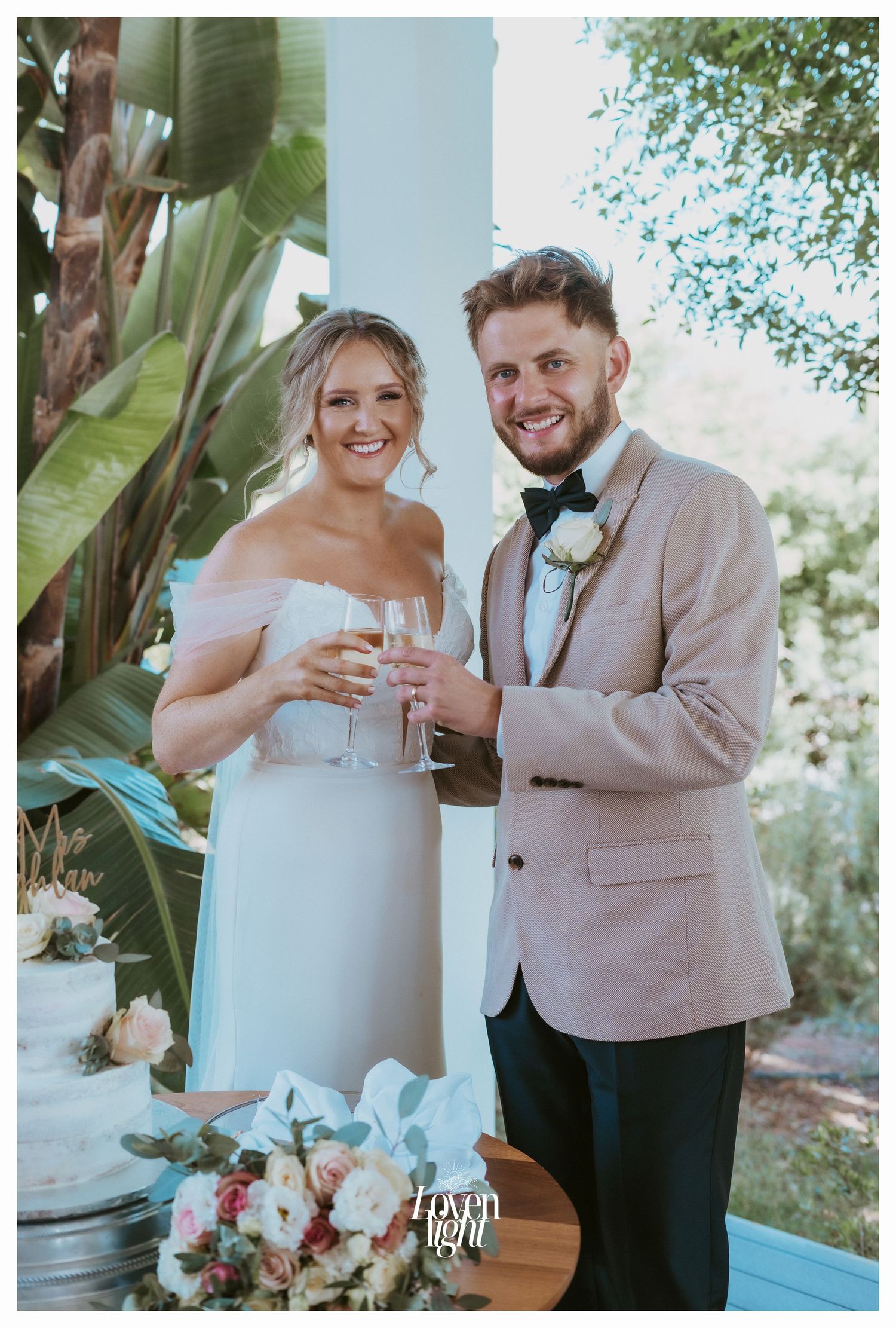 Newlyweds toast with champagne beside their wedding cake in a garden setting.