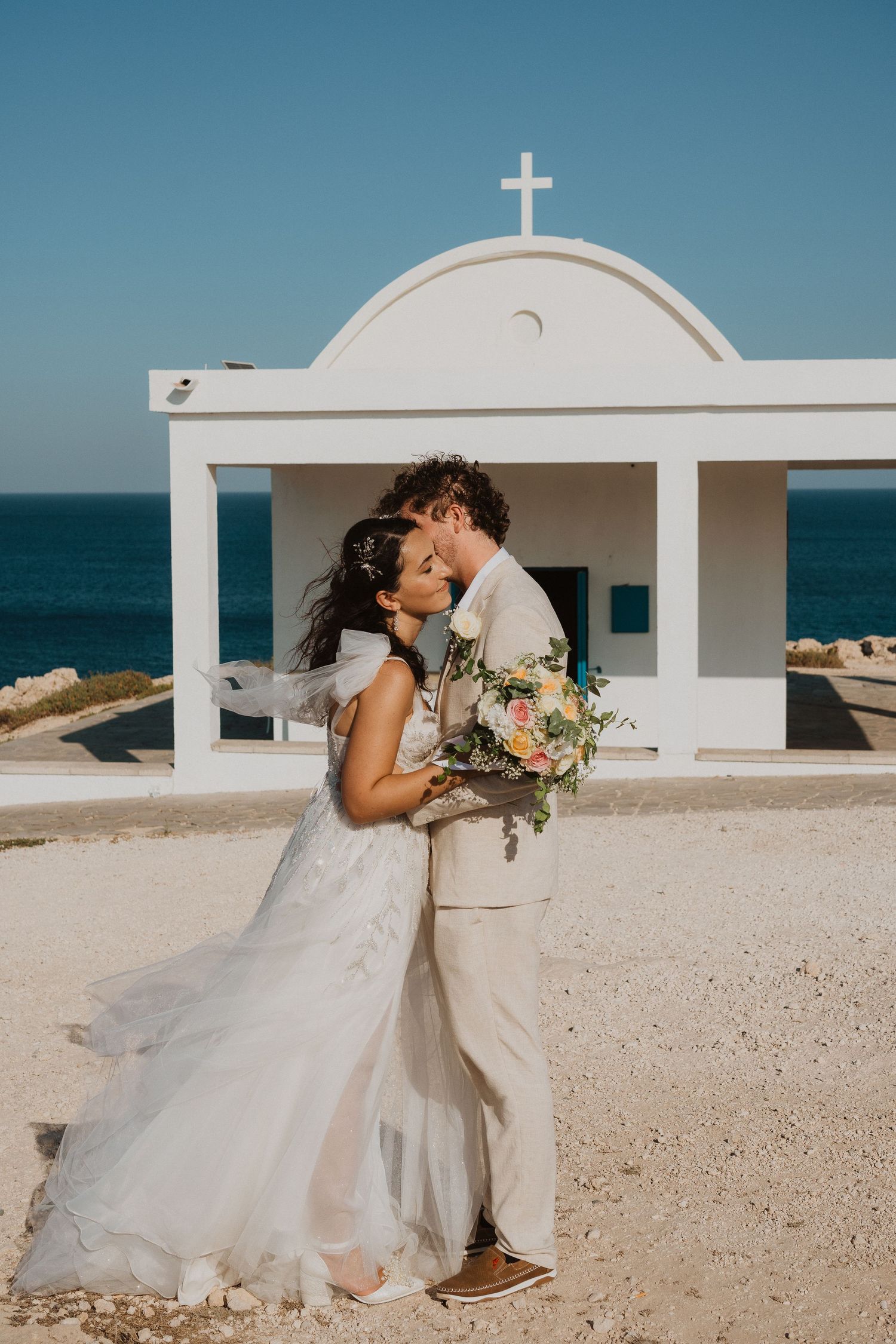 A tender wedding moment captured in front of a seaside chapel with bright blue skies.