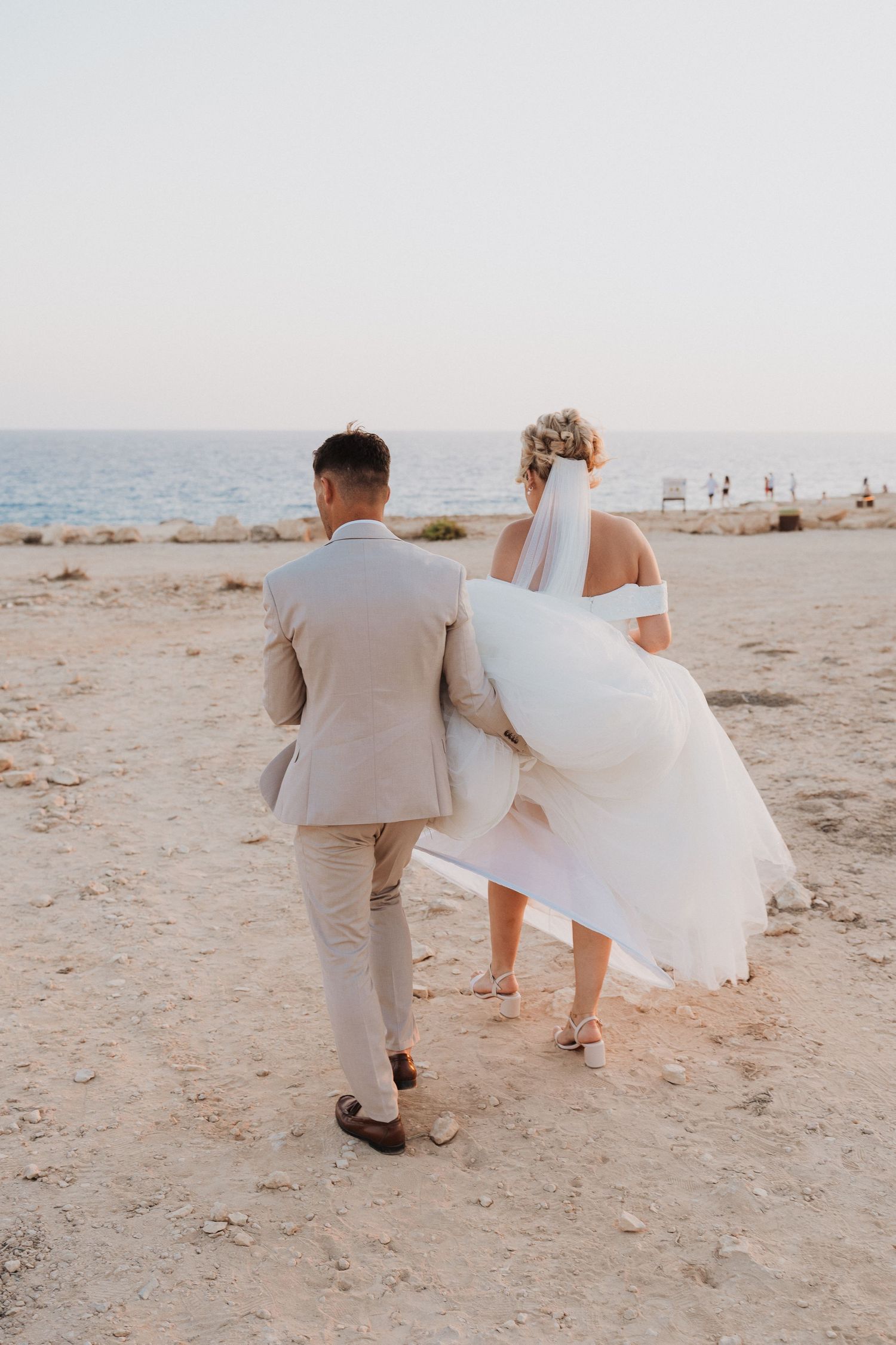 A romantic beach wedding couple walking together on sandy shore with her white dress flowing in the breeze.