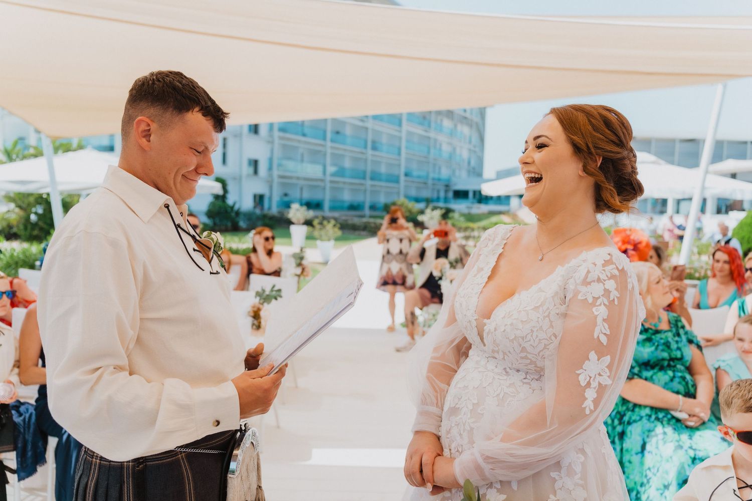 A couple shares a joyful moment during their beach wedding ceremony while exchanging vows under a white canopy.