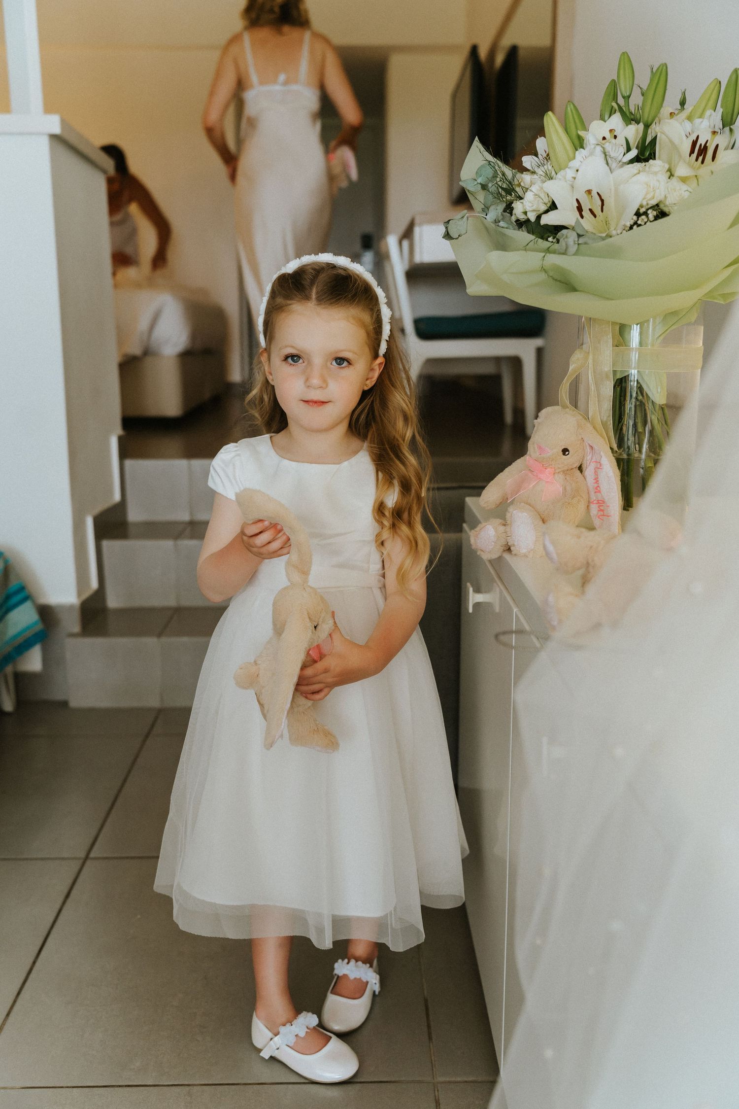 Young attendant in white dress holding stuffed bunny near floral arrangement.