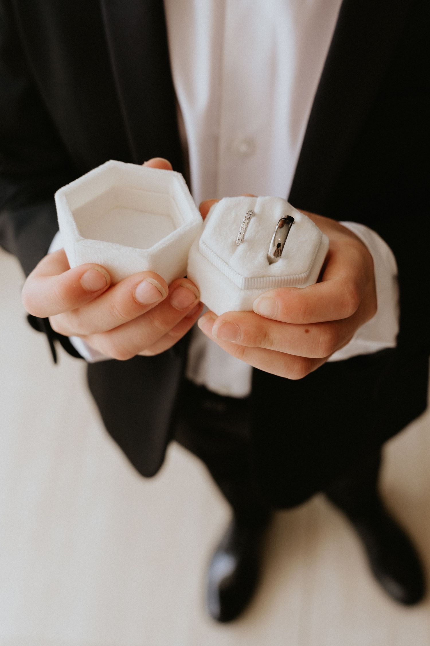 Hands holding a white hexagonal ring box against a black suit.