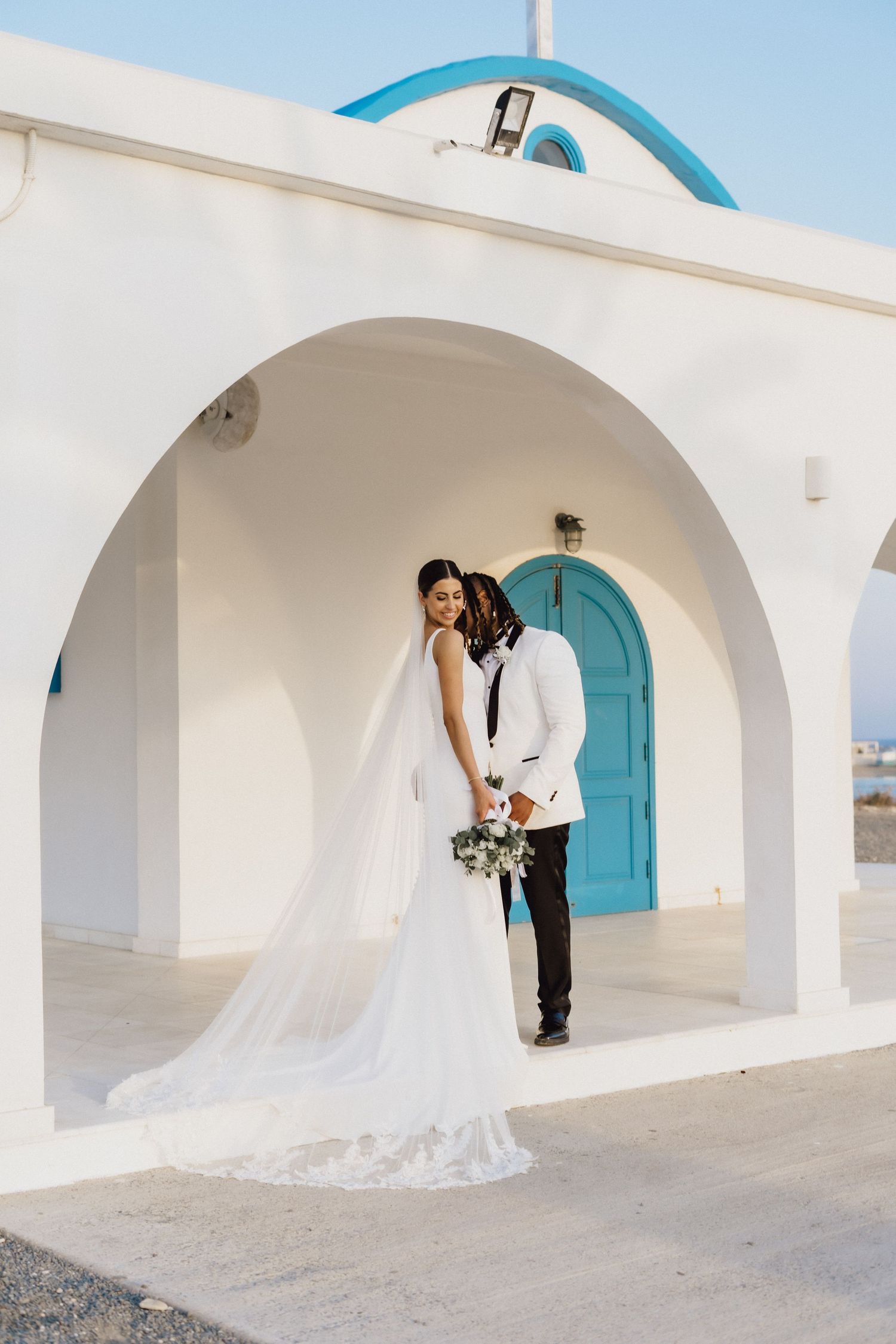 A wedding couple poses under white arches with a stunning turquoise door and flowing veil.