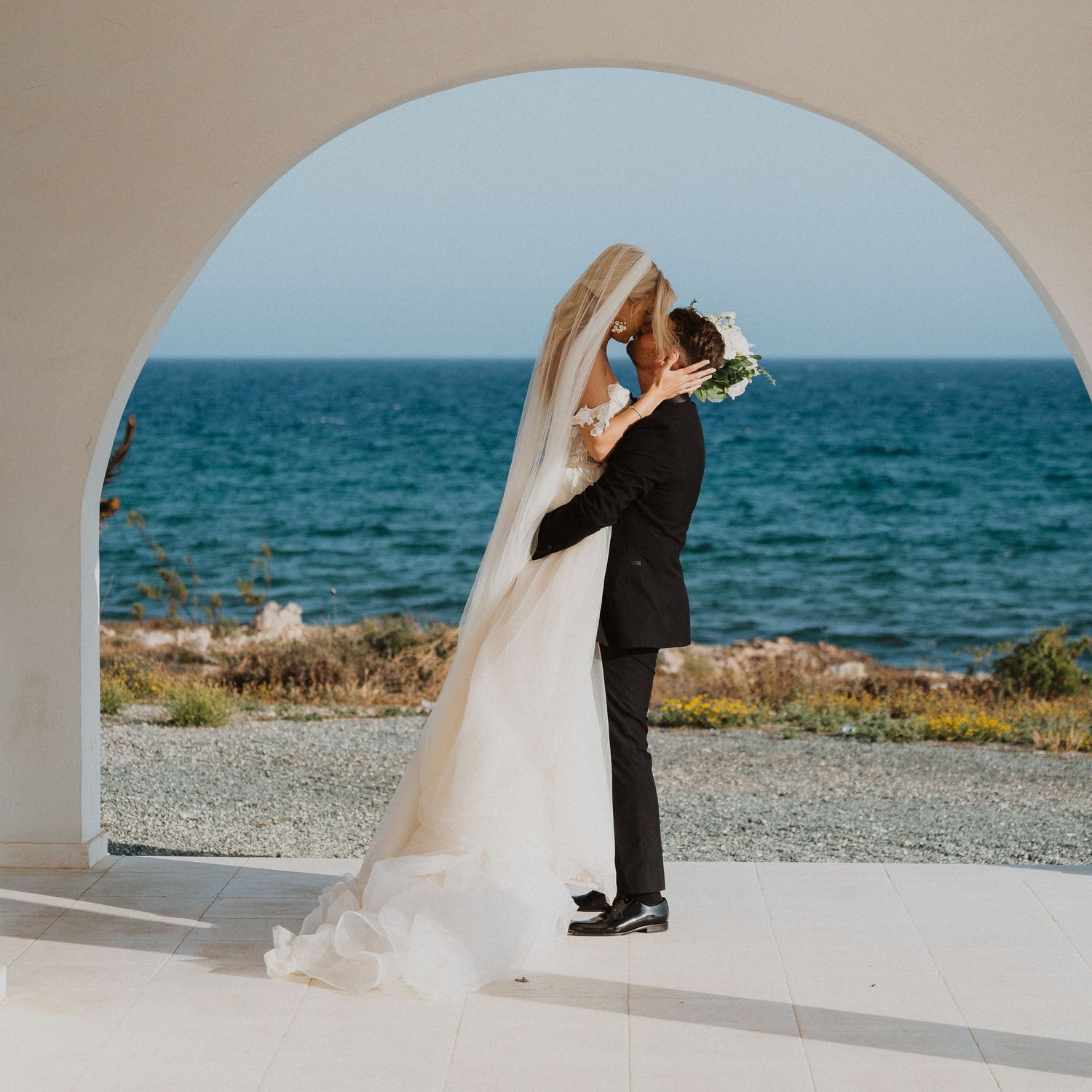 Newlyweds share an intimate moment under an archway by the Mediterranean Sea.