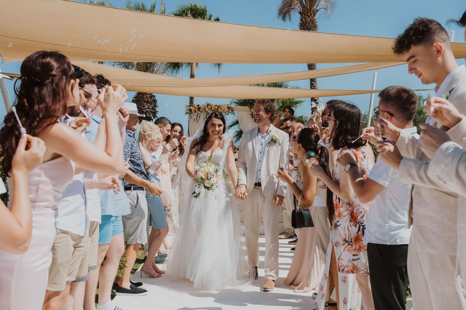 Guests blow bubbles as newlyweds walk down aisle at beach wedding under canvas shades.