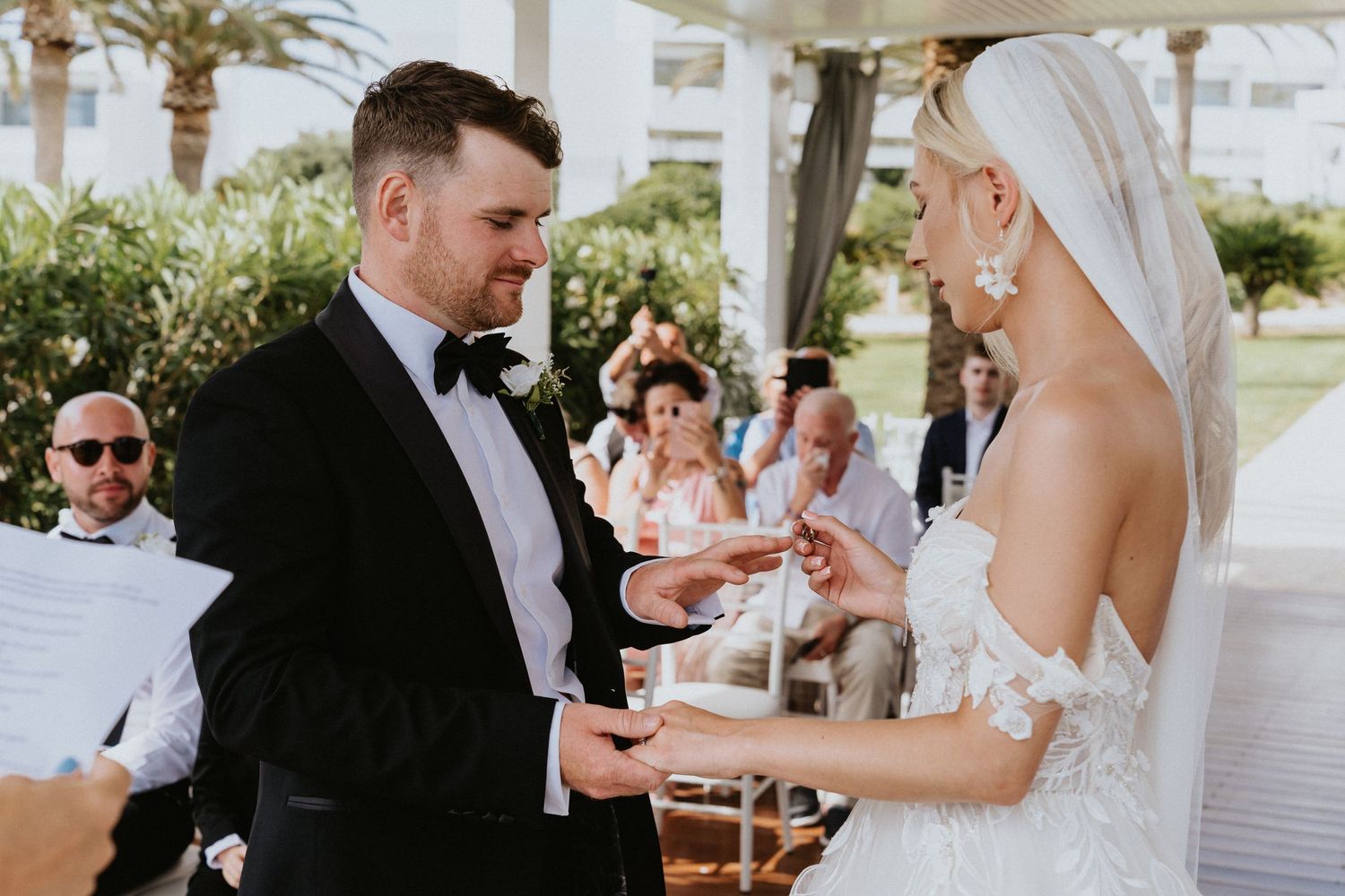 A wedding ceremony shows exchange of rings during an outdoor coastal celebration.